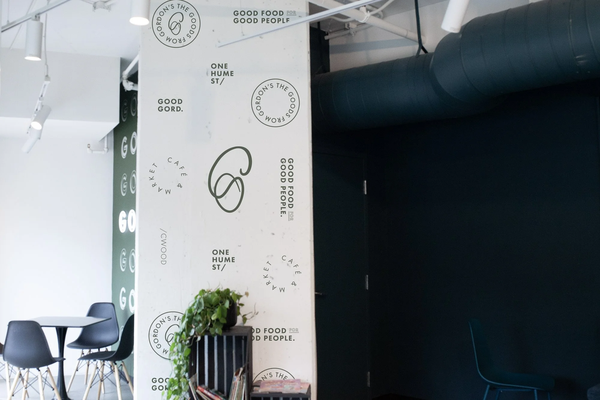 Interior of a modern cafe with a white wall featuring multiple logos and slogans for 'Gordon's' including phrases like 'Good Food for Good People' and 'One Hume St.' There are black chairs with wooden legs and a small black table, a potted green plan