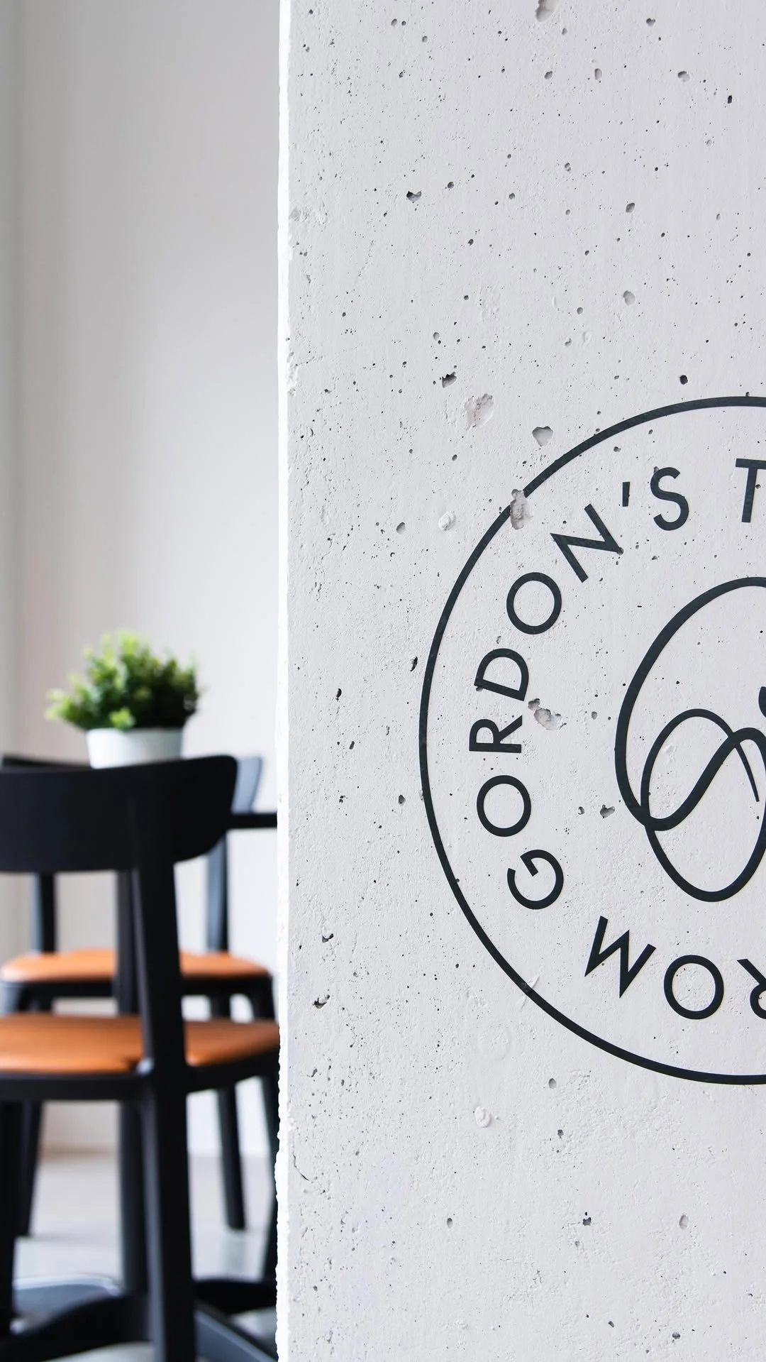 A close-up of a white textured concrete wall featuring a circular logo that reads 'GOOD REST' with an abstract design in the center. In the background, part of a black chair with wooden armrests and a small potted plant on a table are visible, indica