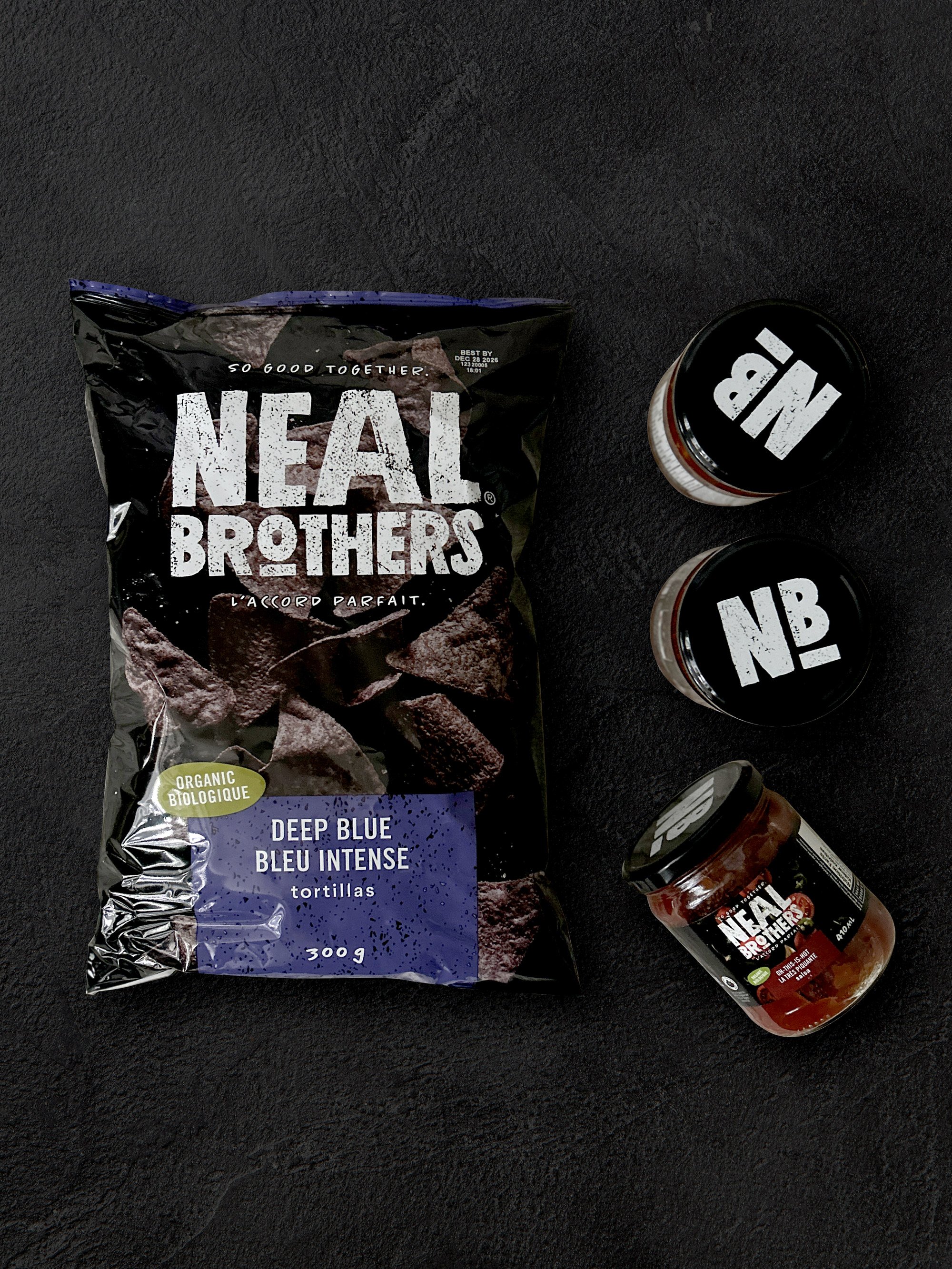 A bag of Neal Brothers organic deep blue tortillas and three jars of Neal Brothers salsa on a black surface.