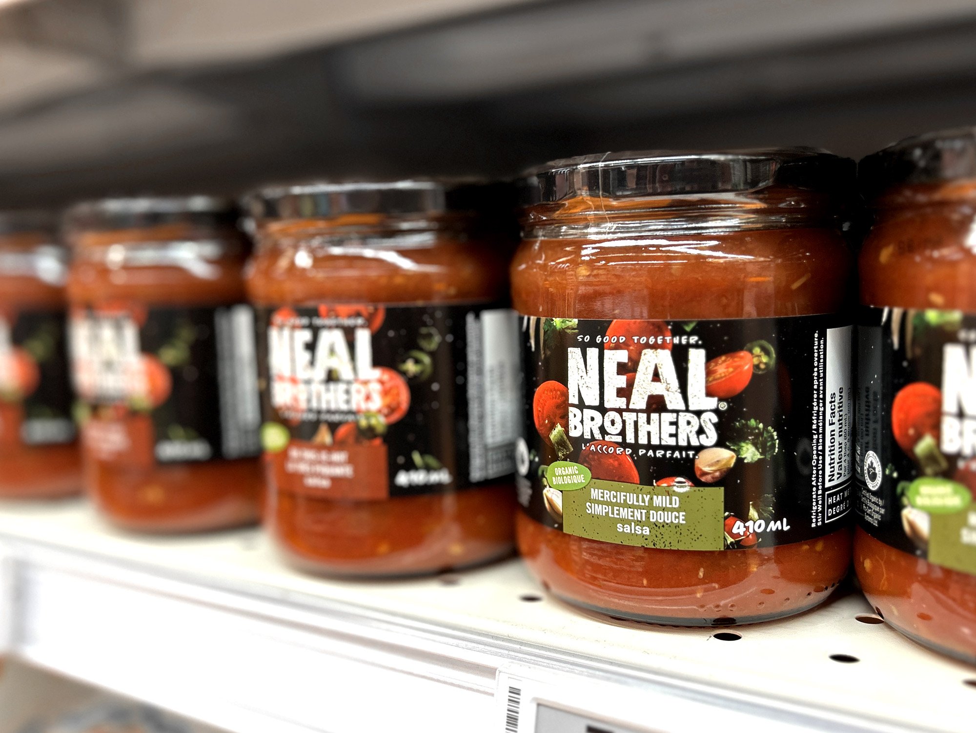 Multiple jars of Neal Brothers organic mild salsa on store shelf. The jar labels are black with images of tomatoes and mentions the salsa is simple and mild. 