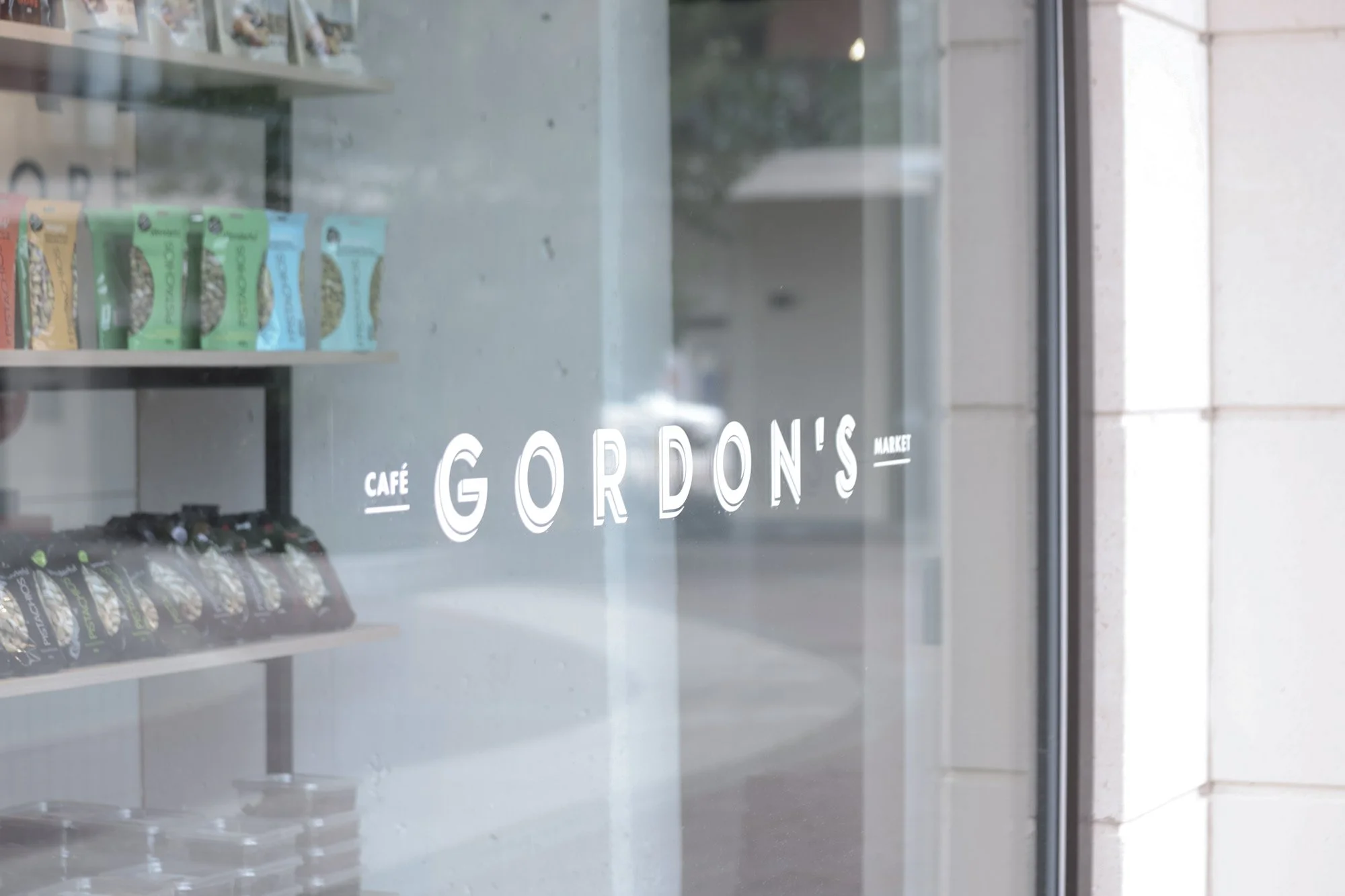 The image shows a glass storefront window with the text 'Gordon's' and 'Café Market'. Inside, there are shelves with snack items or packaged goods, and the scene appears to be a modern, clean retail or café space.