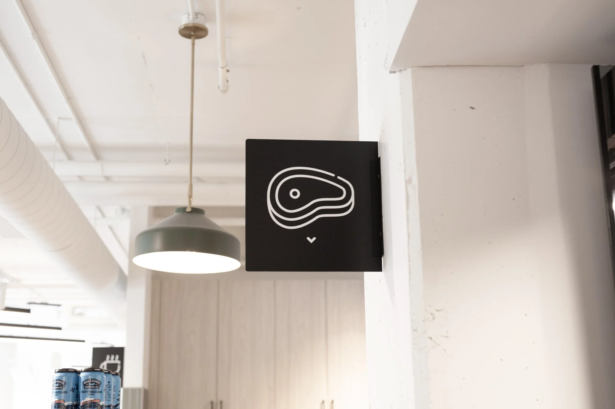 Grocery store wayfinding, a black blade sign featuring a white outline of a piece of meat, possibly steak, on a white surface in a bright, modern interior with white walls, ceiling, and ventilation duct, and a hanging green lamp in the background.