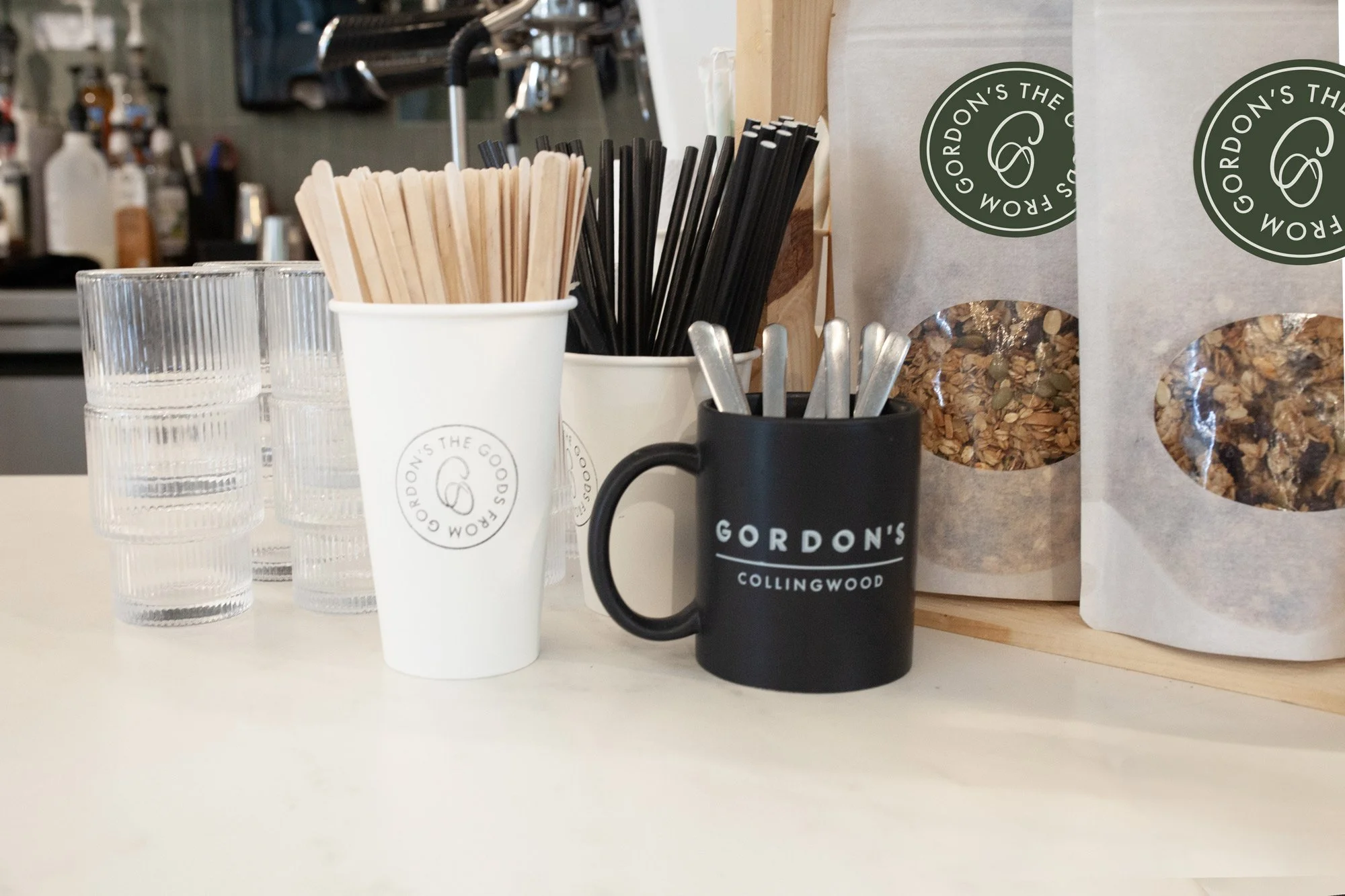 Inside a café, several clear glasses are on a white counter, along with a white mug and a black mug marked 'Gordon's Collingwood'. There are containers with wooden and black straws, and paper bags of granola with green circular labels. The background