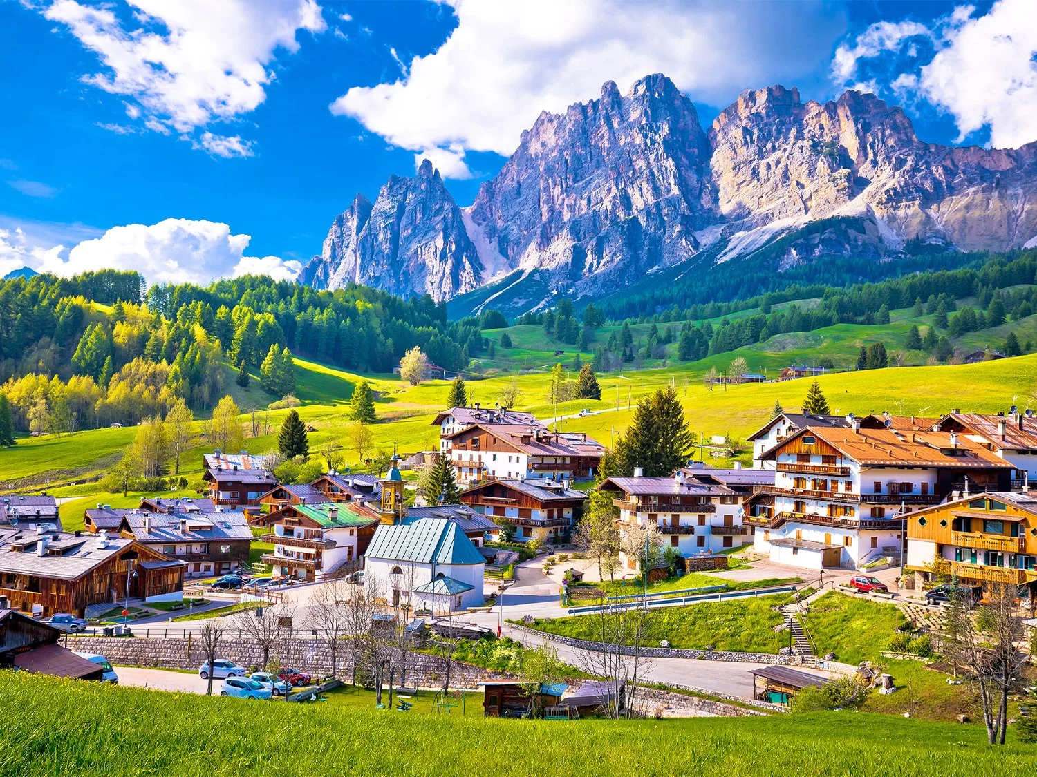 A mountain village with houses and buildings at the base of towering, rocky mountains under a blue sky with clouds.