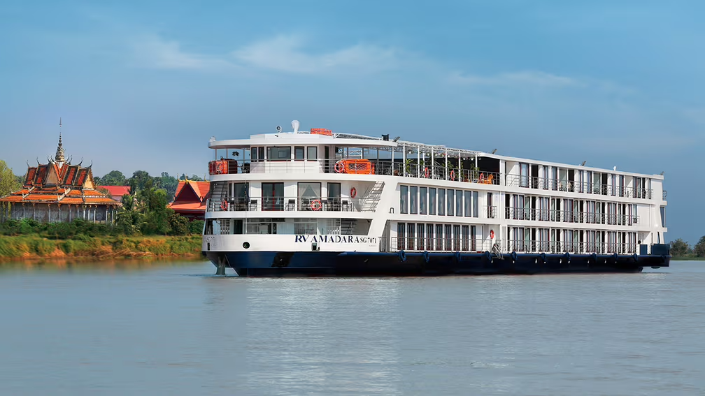 Large white and black river cruise ship with multiple decks sailing on calm water near a riverside with traditional Thai-style buildings and green trees.