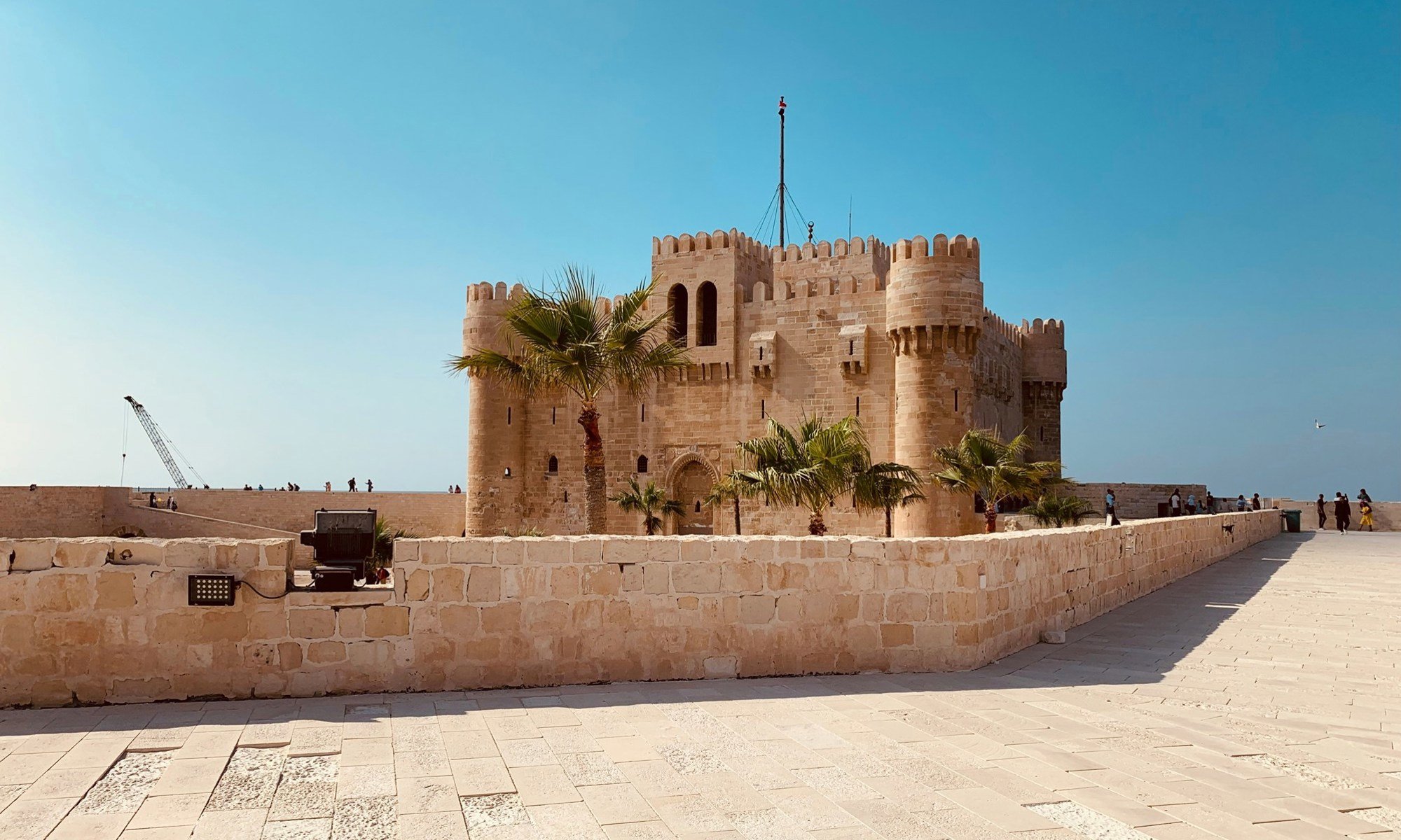 A historic stone fortress with cylindrical towers and crenellated walls, surrounded by palm trees, against a clear blue sky. People are walking along a paved pathway near the fortress.