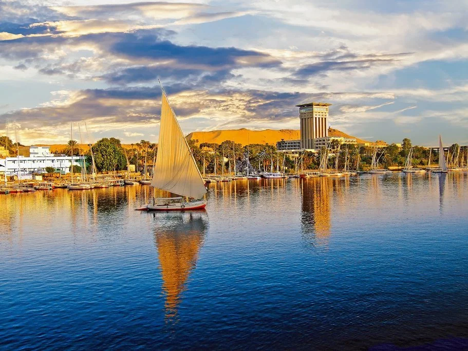 A sailboat with a beige sail on calm water with reflections, a city skyline in the background, and a golden hill under a cloudy sky during sunset.