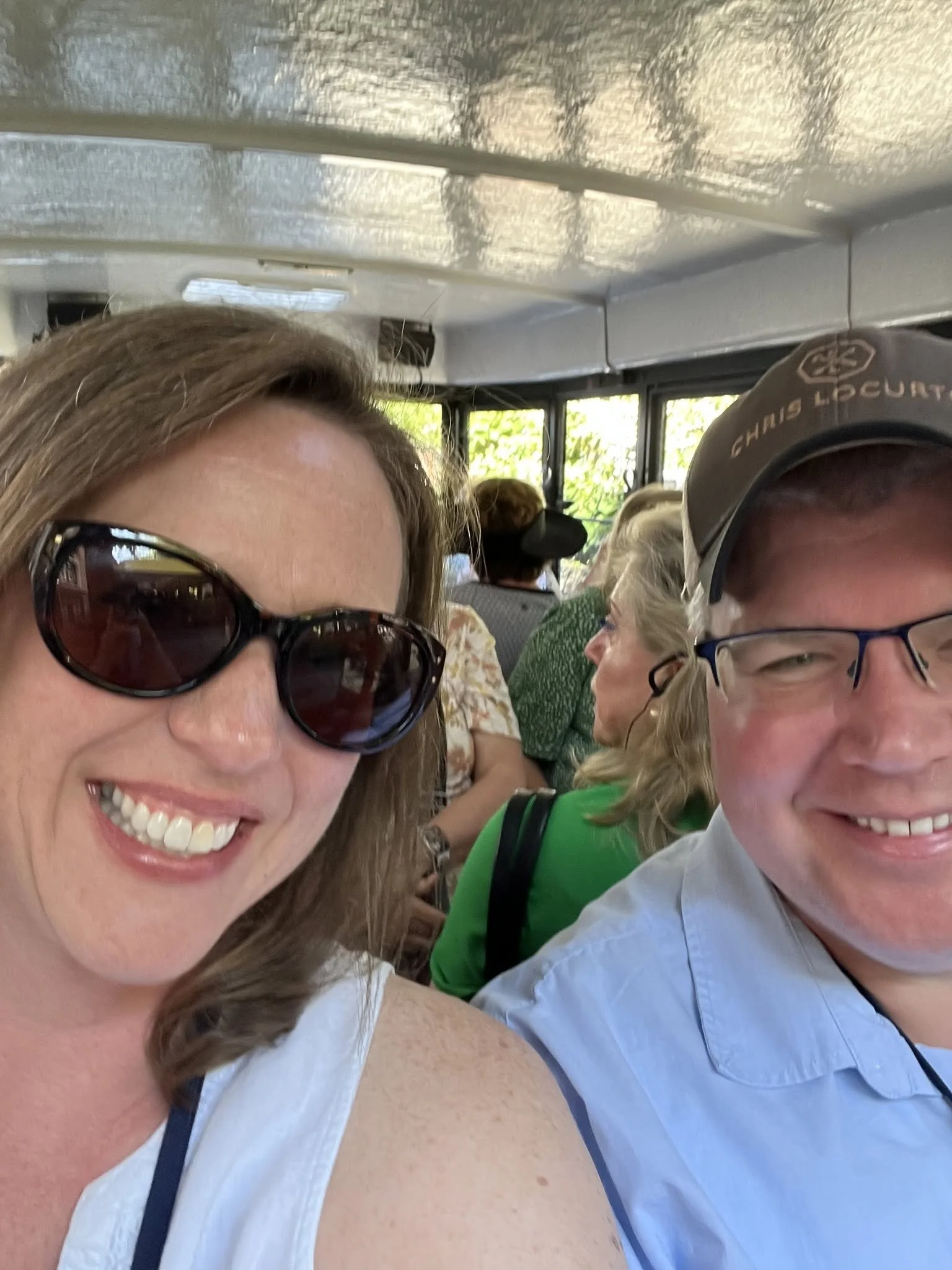 Close-up of smiling woman with sunglasses and woman in baseball cap on a bus with other passengers visible in the background.