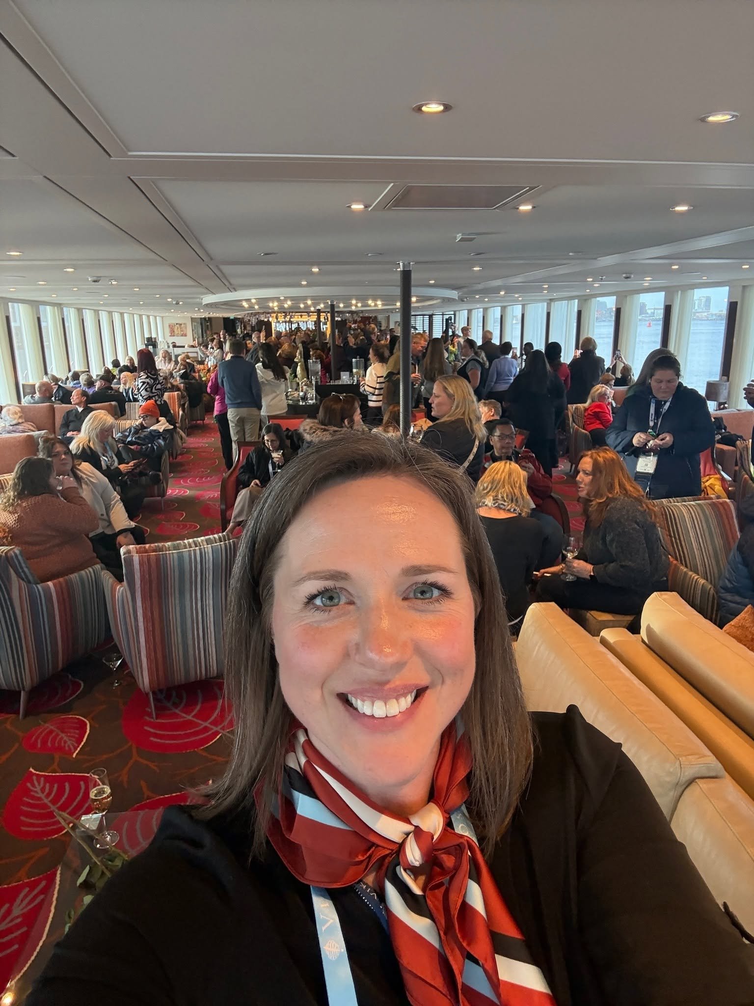 A smiling woman taking a selfie in a busy, well-lit lounge with many people, large windows, and river view.