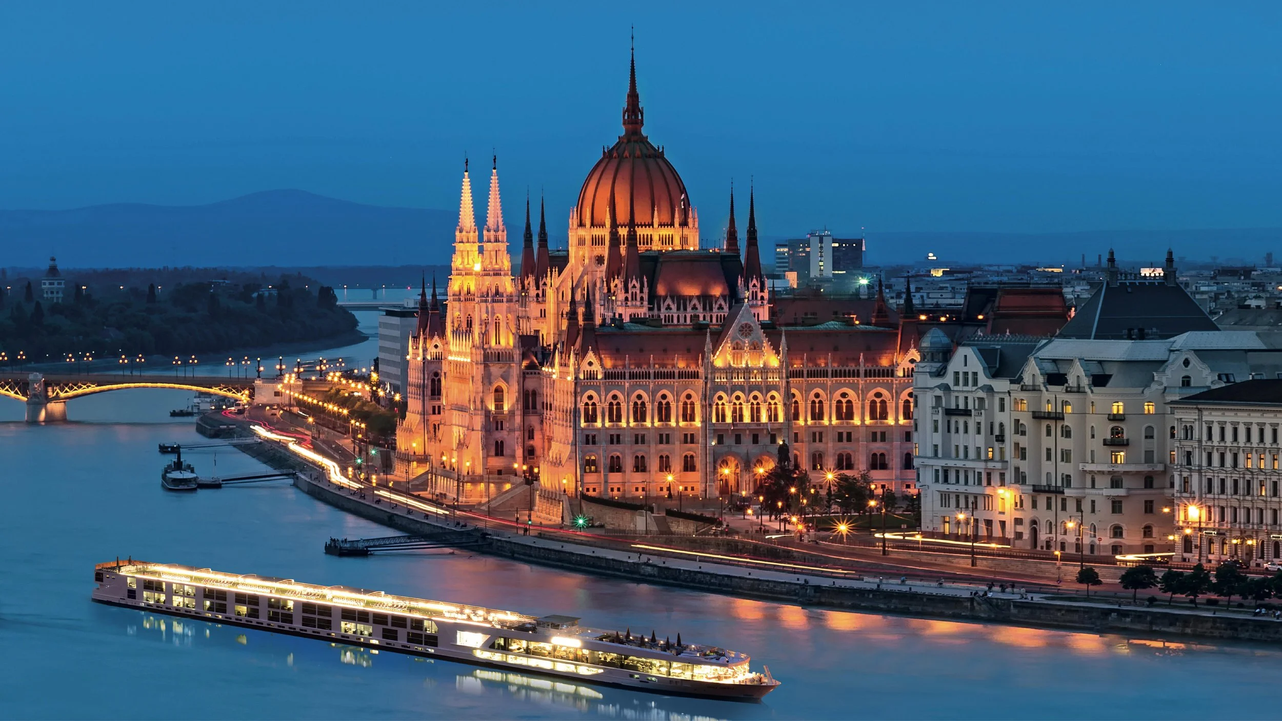 Night view of the Hungarian Parliament Building illuminated by lights along the riverbank with a boat cruising on the Danube River in Budapest, Hungary.