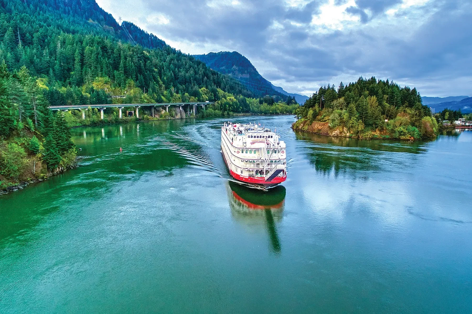 A large white and red cruise ship sailing on a river surrounded by lush green forests and hills, with a bridge visible in the background under a cloudy sky.