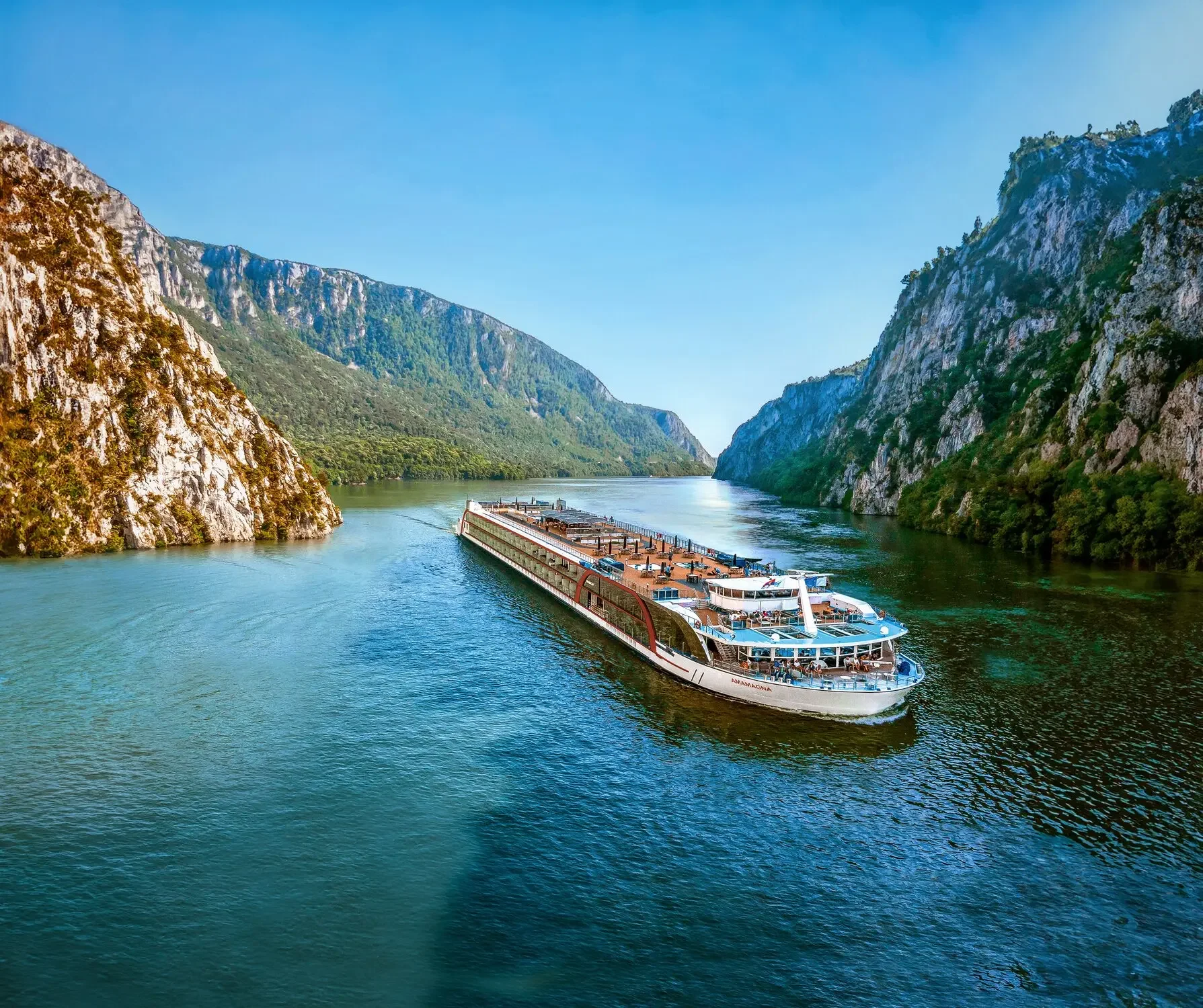 A large cruise ship sailing through a narrow river gorge with steep, rocky cliffs on either side and lush green vegetation, under a clear blue sky.