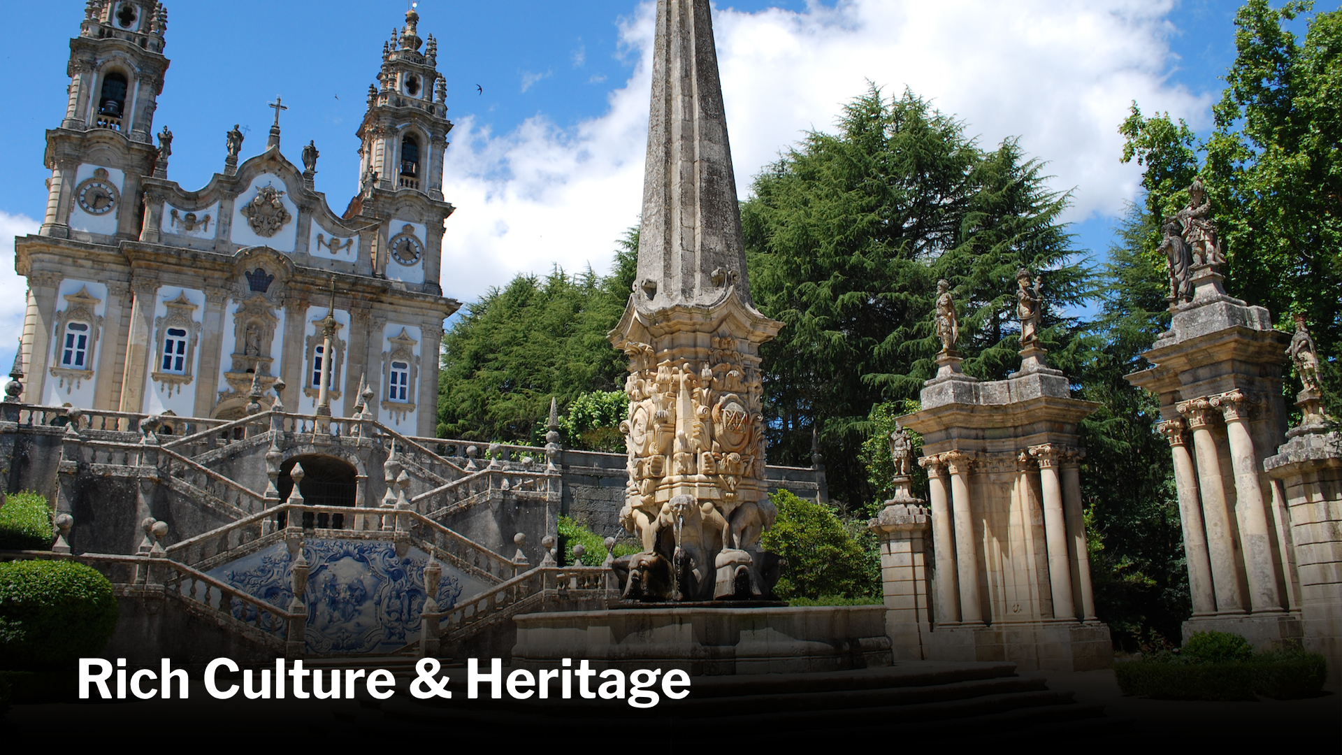 Historic European church with ornate architecture, surrounded by a stone fence, statues, and lush green trees under a partly cloudy sky.