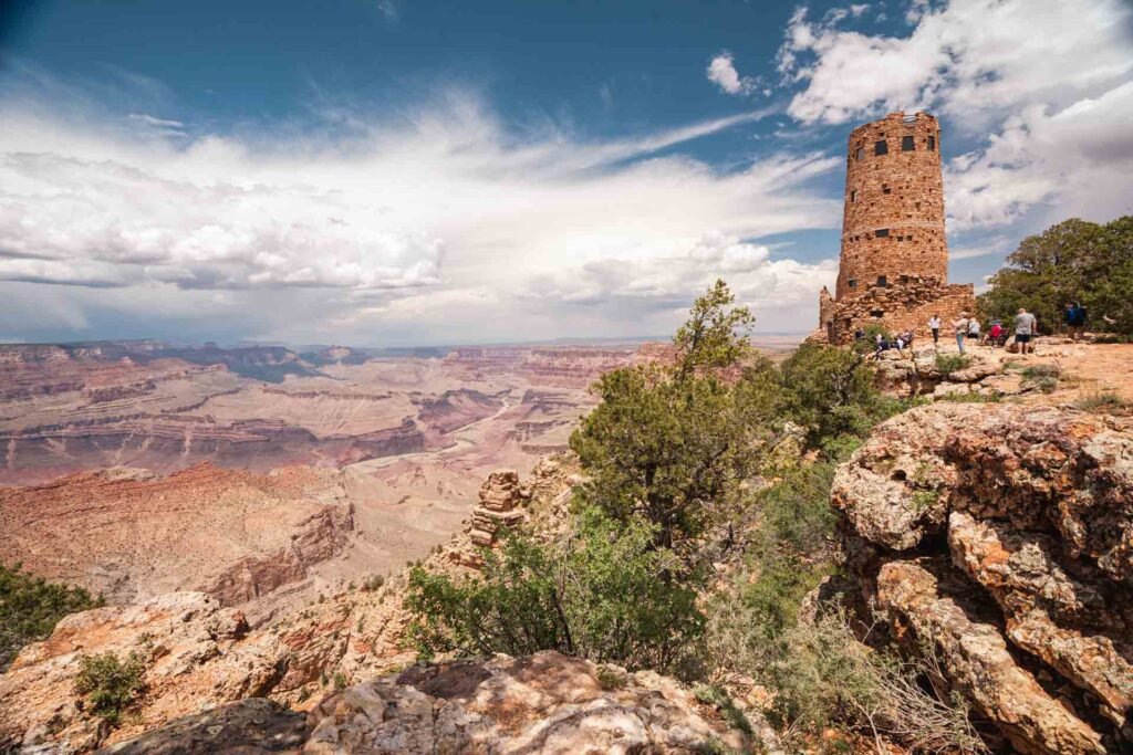A view from the Grand Canyon showing layered rock formations, a tall stone tower, and several visitors exploring the area under partly cloudy skies.