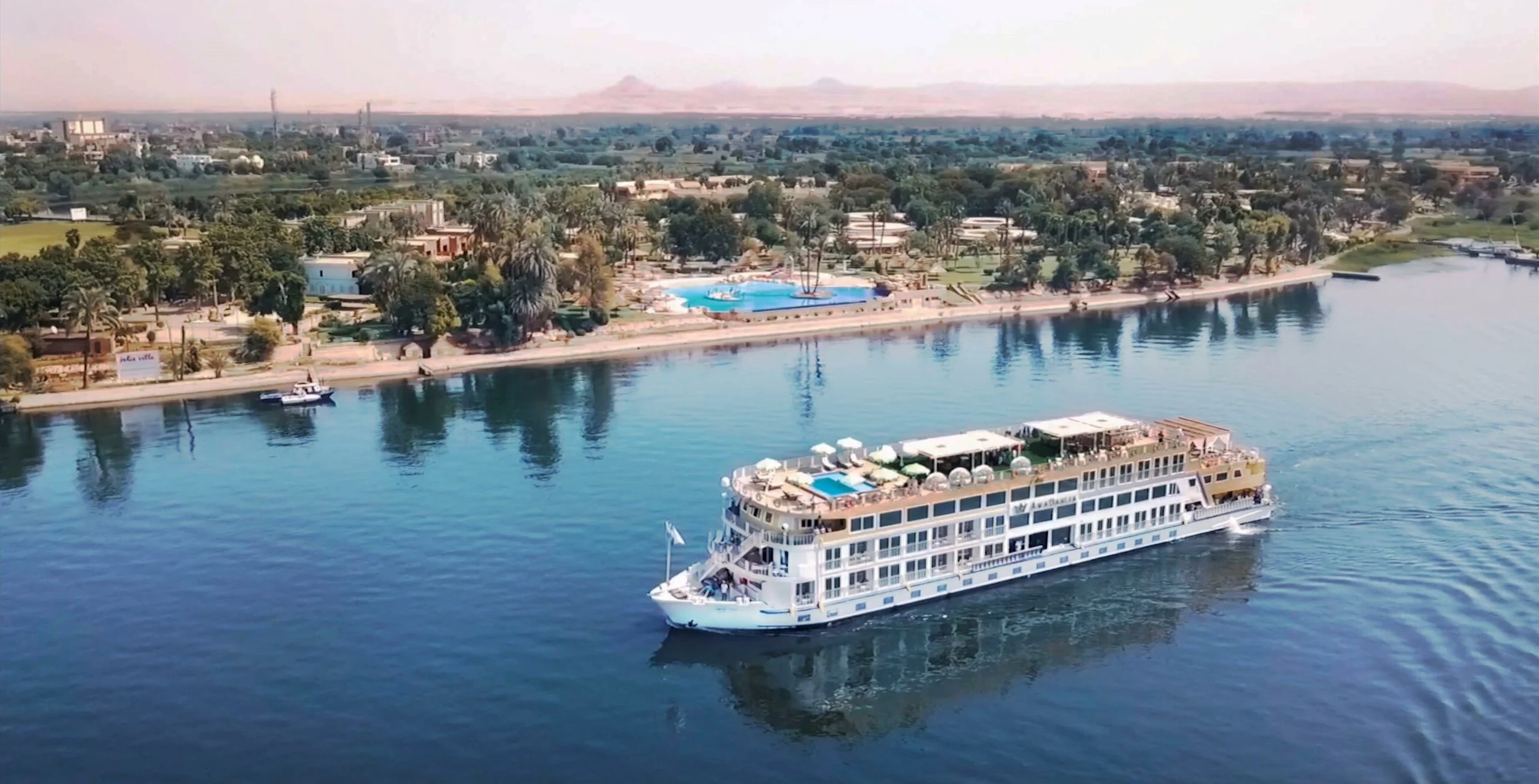 A large white cruise ship with multiple decks, a swimming pool on the upper deck, and outdoor seating, sailing on a calm river near a landscaped park with trees, a swimming pool, and buildings in the background.