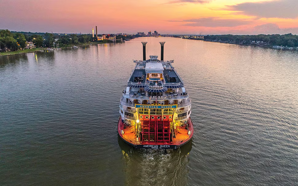 A large passenger cruise ship with the name 'American Queen' is sailing on a river during sunset, with the city skyline in the background.