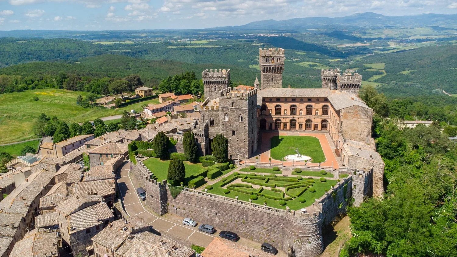 Aerial view of a medieval castle with towers and a courtyard, surrounded by a village and green landscape, under a partly cloudy sky.