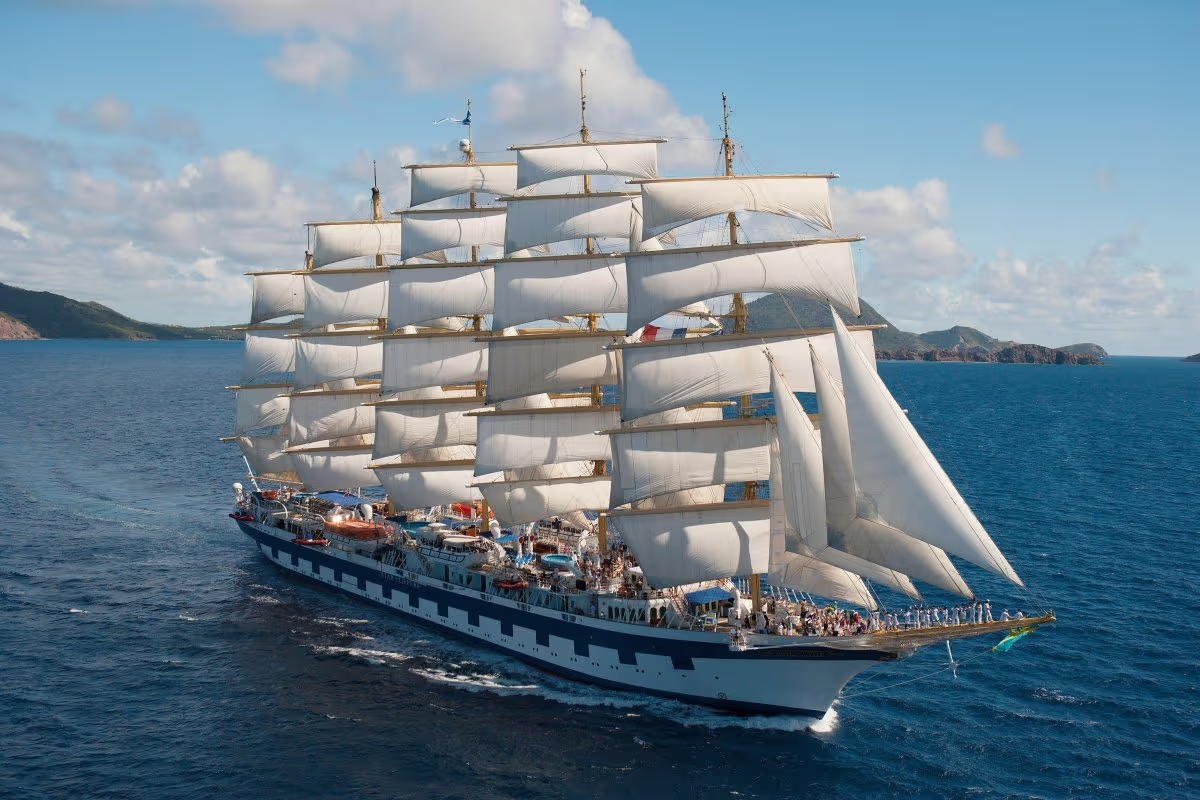 A large sailing ship with multiple white sails on the blue ocean, with land and hills in the background under a partly cloudy sky.