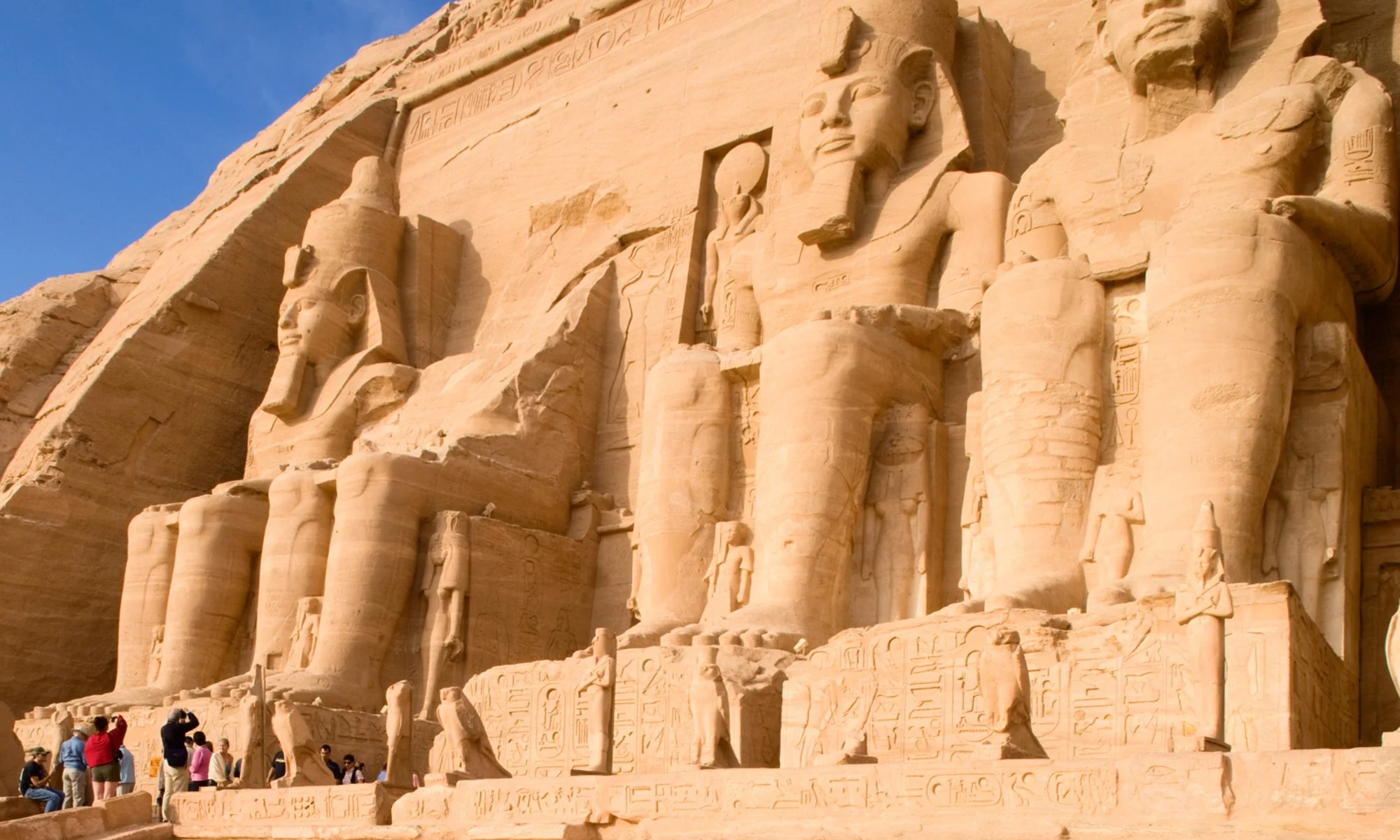 The image shows the ancient Egyptian temple of Abu Simbel with large carved rock statues of Pharaoh Ramses II on its facade. Tourists are gathered in front of the temple, taking photos. The sky is clear and blue.