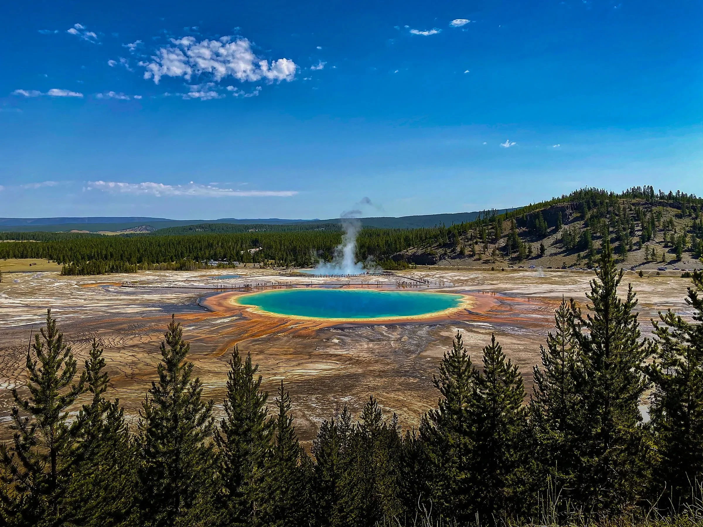 A geothermal hot spring with colorful mineral deposits, steam rising from the water, surrounded by trees and a hilly landscape under a blue sky with scattered clouds.