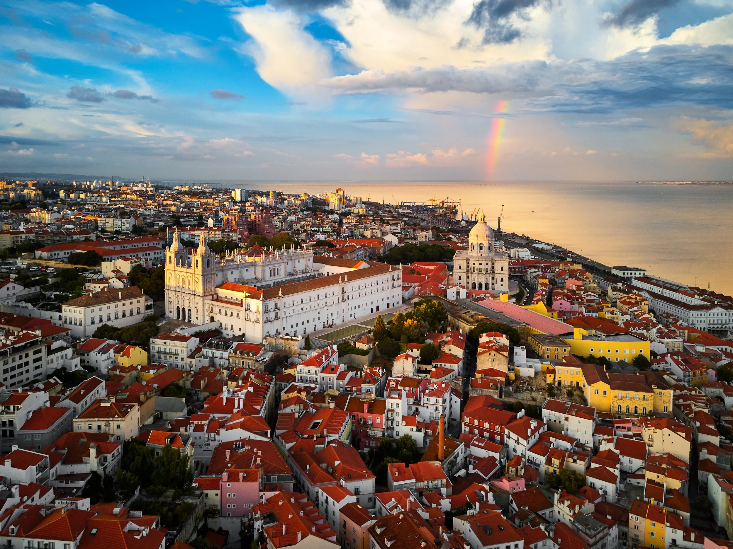 A cityscape showing a mix of historic and modern buildings with red-tiled rooftops, near a body of water, with a rainbow in a partly cloudy sky at sunset.