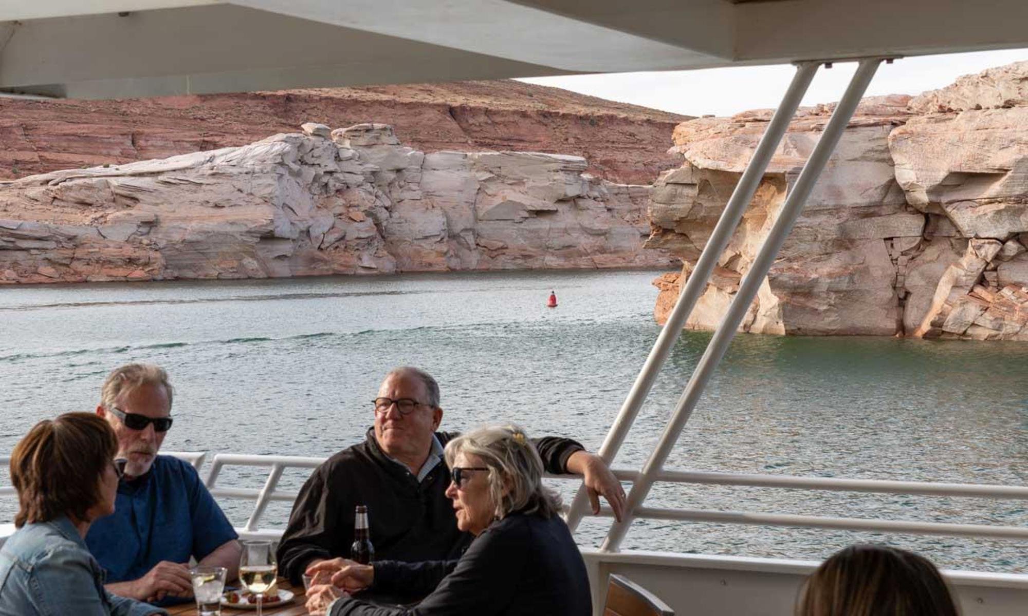 People sitting on a boat enjoying drinks with a view of red rock cliffs and water in the background.