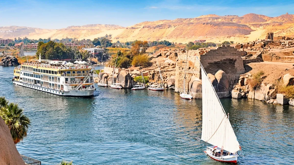 A river scene with a large white cruise boat, several smaller sailboats, and rocky, desert-like hills in the background.