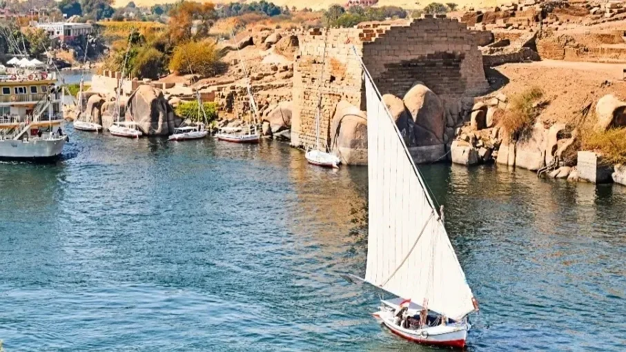 A sailboat with a white sail moving through a body of water next to rocky cliffs. Several other boats are docked nearby along the shoreline, with small buildings and vegetation in the background.