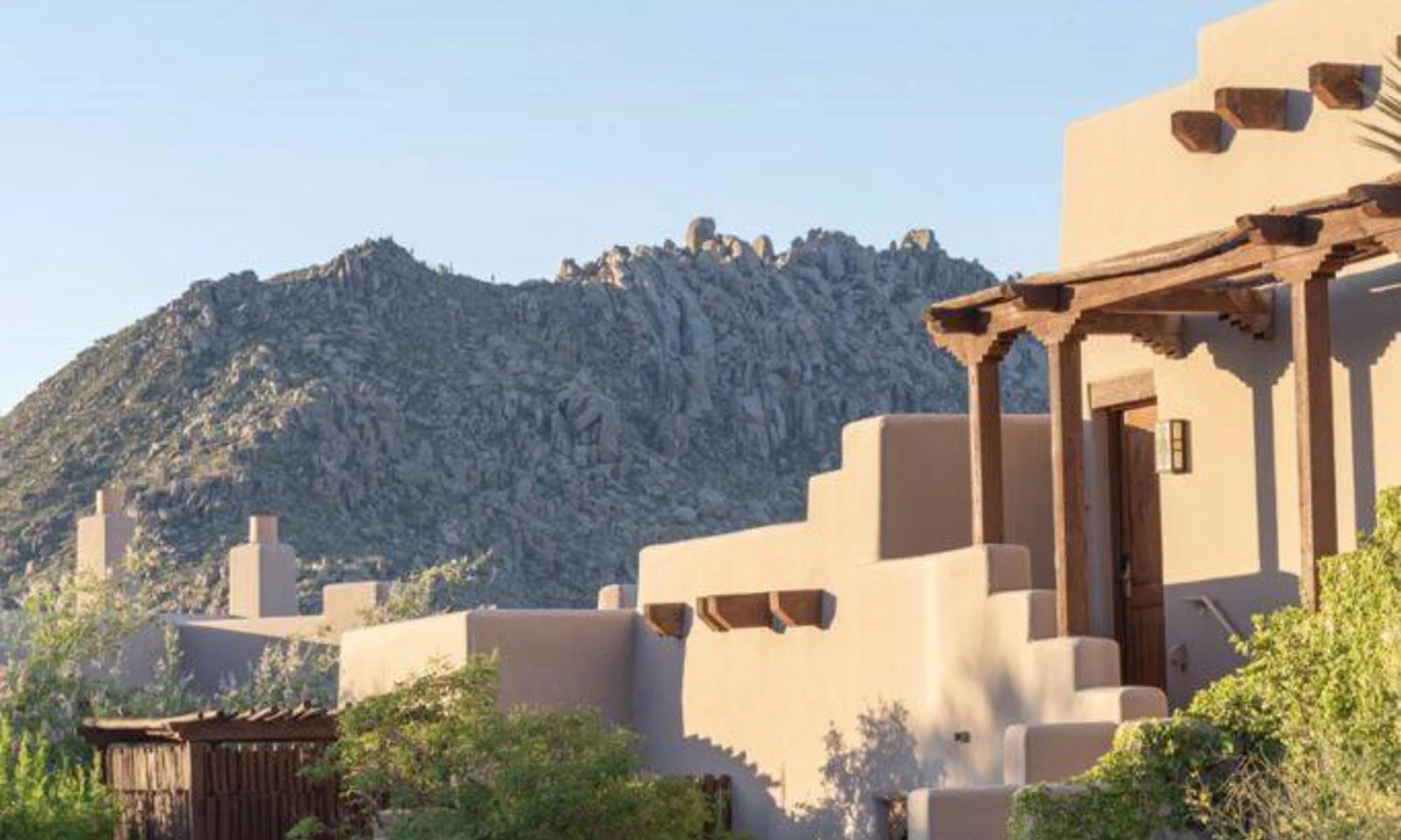 A Southwestern style house with stucco walls and wooden beams, with rocky mountains in the background under a clear blue sky.