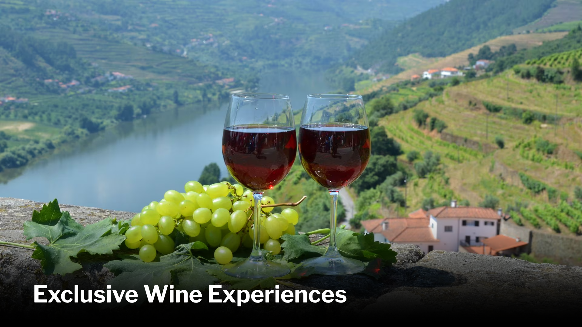 Two glasses of red wine, a cluster of green grapes, and green grape leaves on a stone surface with a scenic river and hillside vineyards in the background.