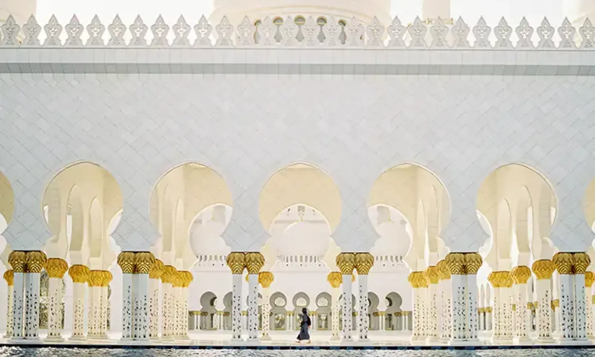 White mosque with arches, topped with golden domes and intricate details, with a person walking along the water in front.