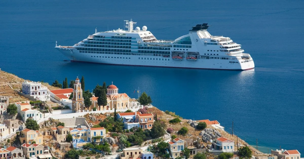 Large white cruise ship sailing near a coastal village with colorful houses and a church with a red dome.