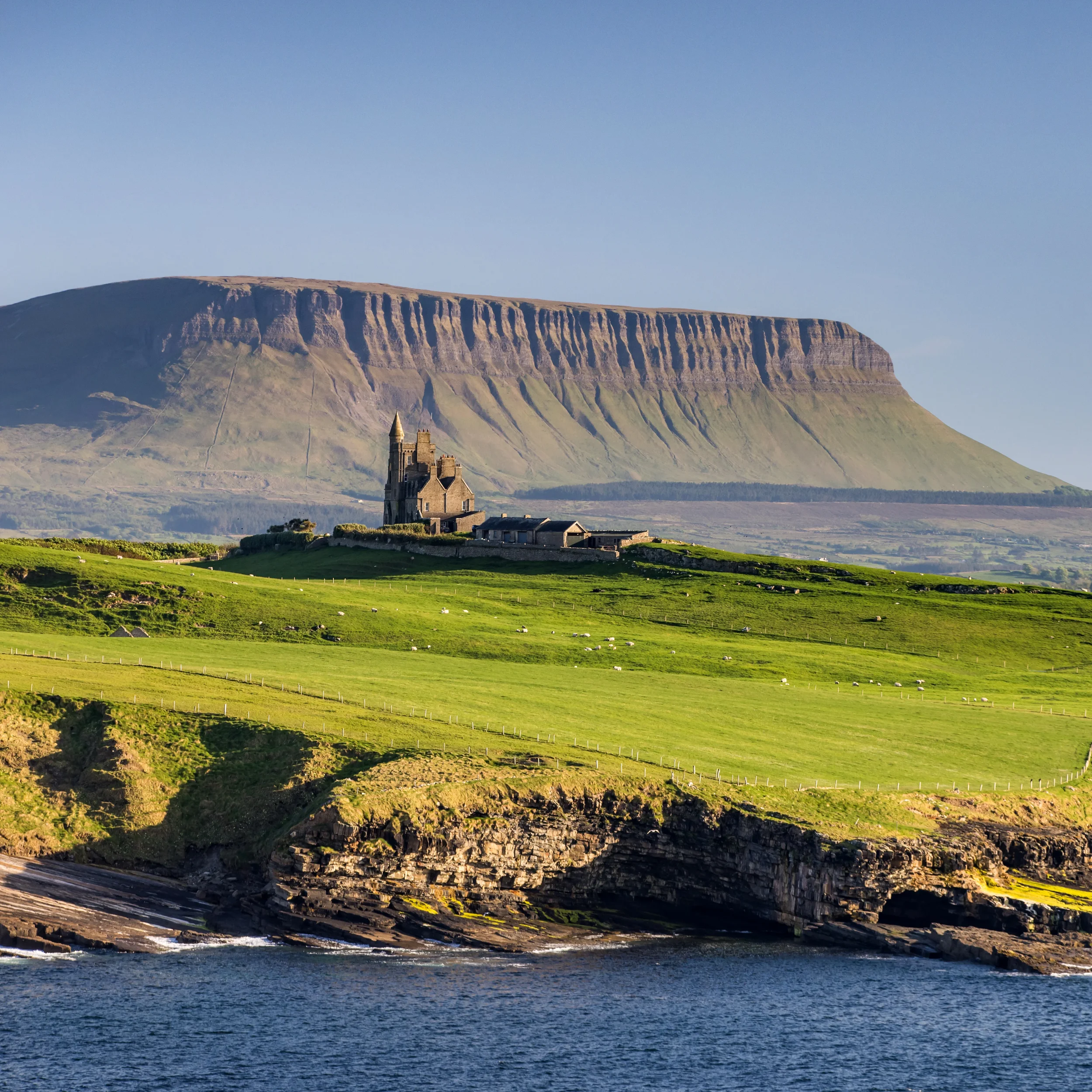 A scenic view of a coastal landscape with a castle on the shore, green hills, cliffs, and a large flat-topped mountain with vertical striations in the background under a clear blue sky.