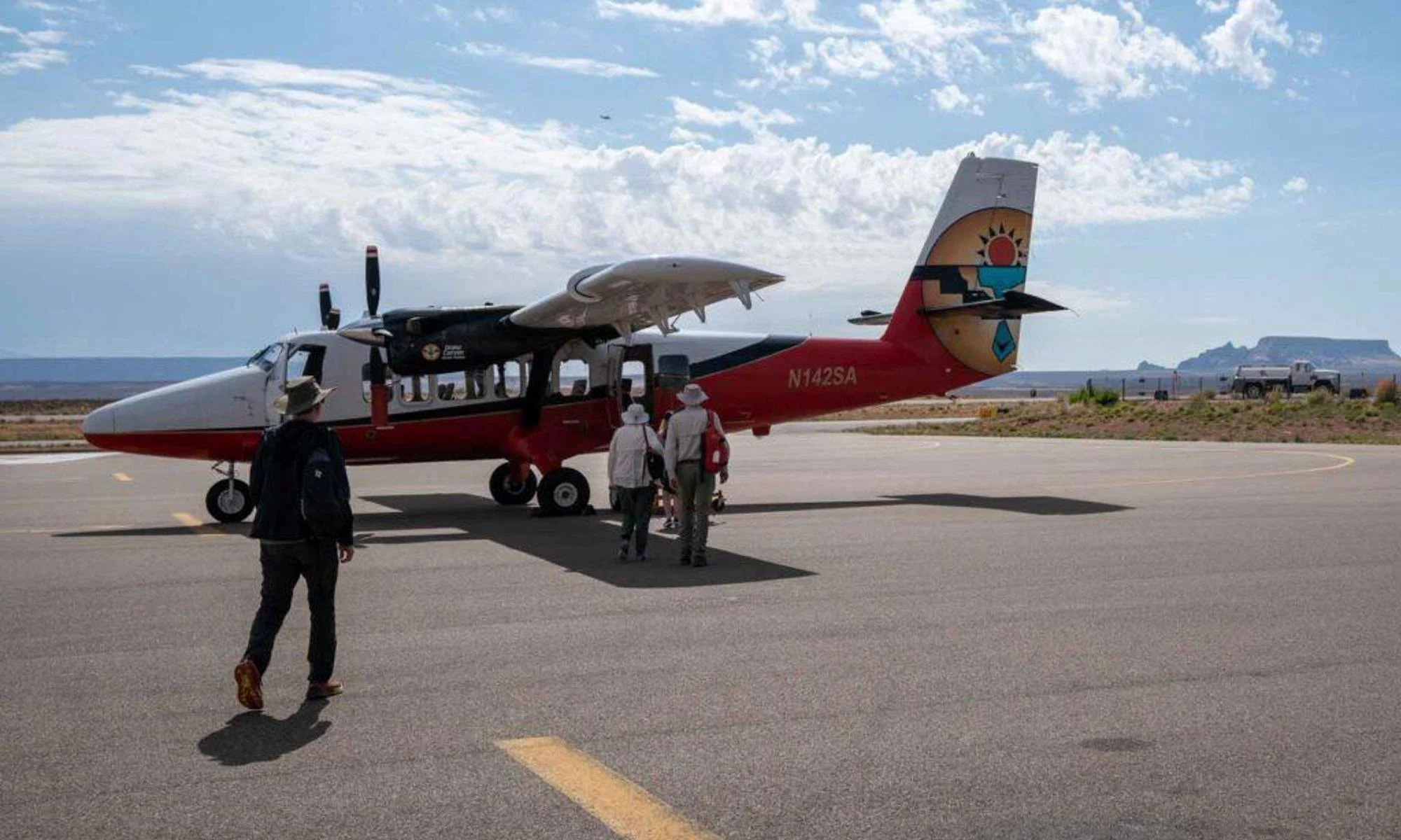 People walking towards a small airplane on an airport tarmac with a cloudy sky and mountains in the background.