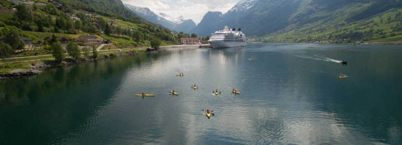A scenic fjord with green hills, mountains, and snow-capped peaks in the background. A large cruise ship is docked near a small town, with several kayakers and boats on the water.