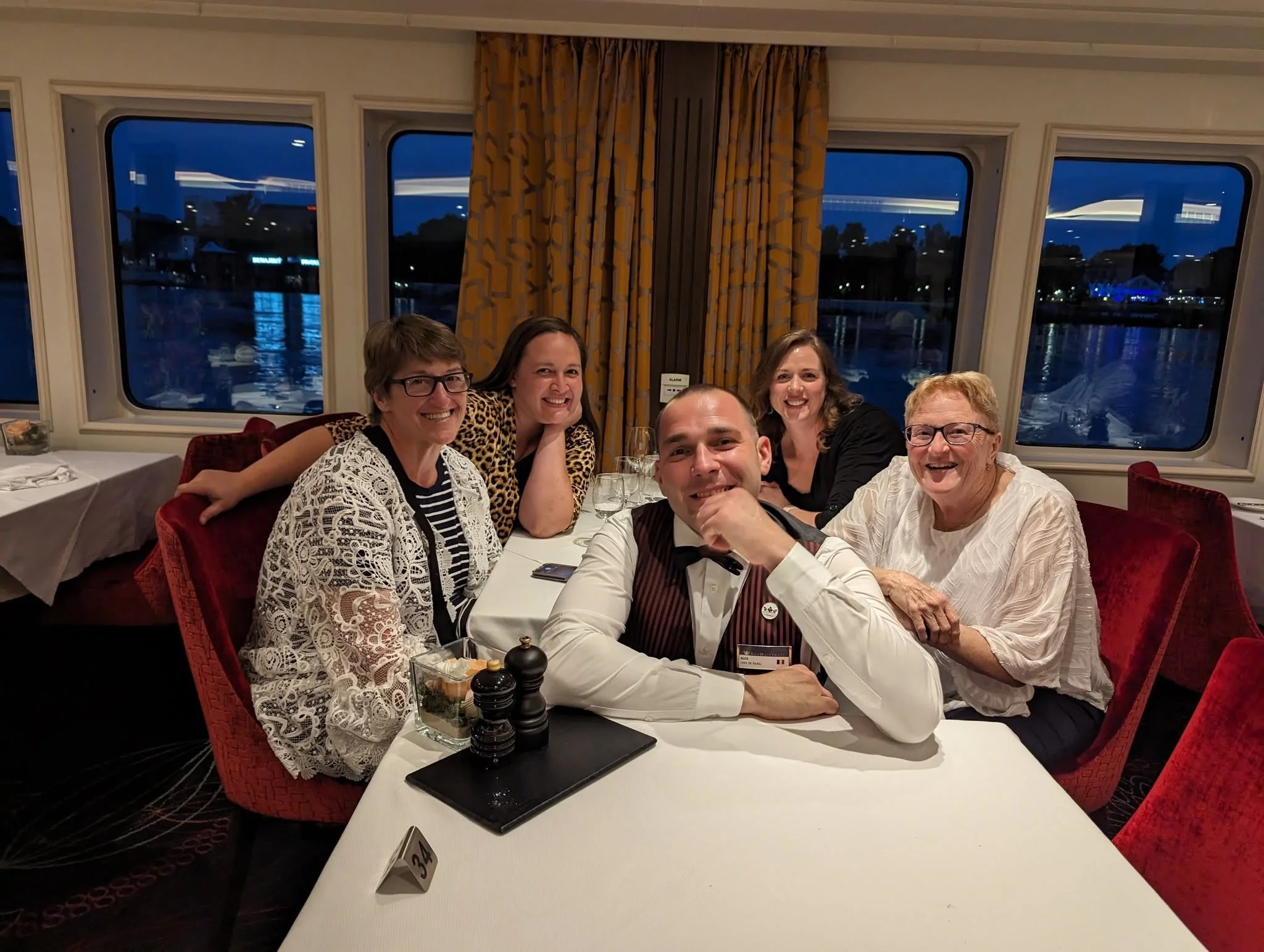 Group of six people sitting at a dining table on a cruise ship, smiling, with a night view of water and buildings outside the windows.