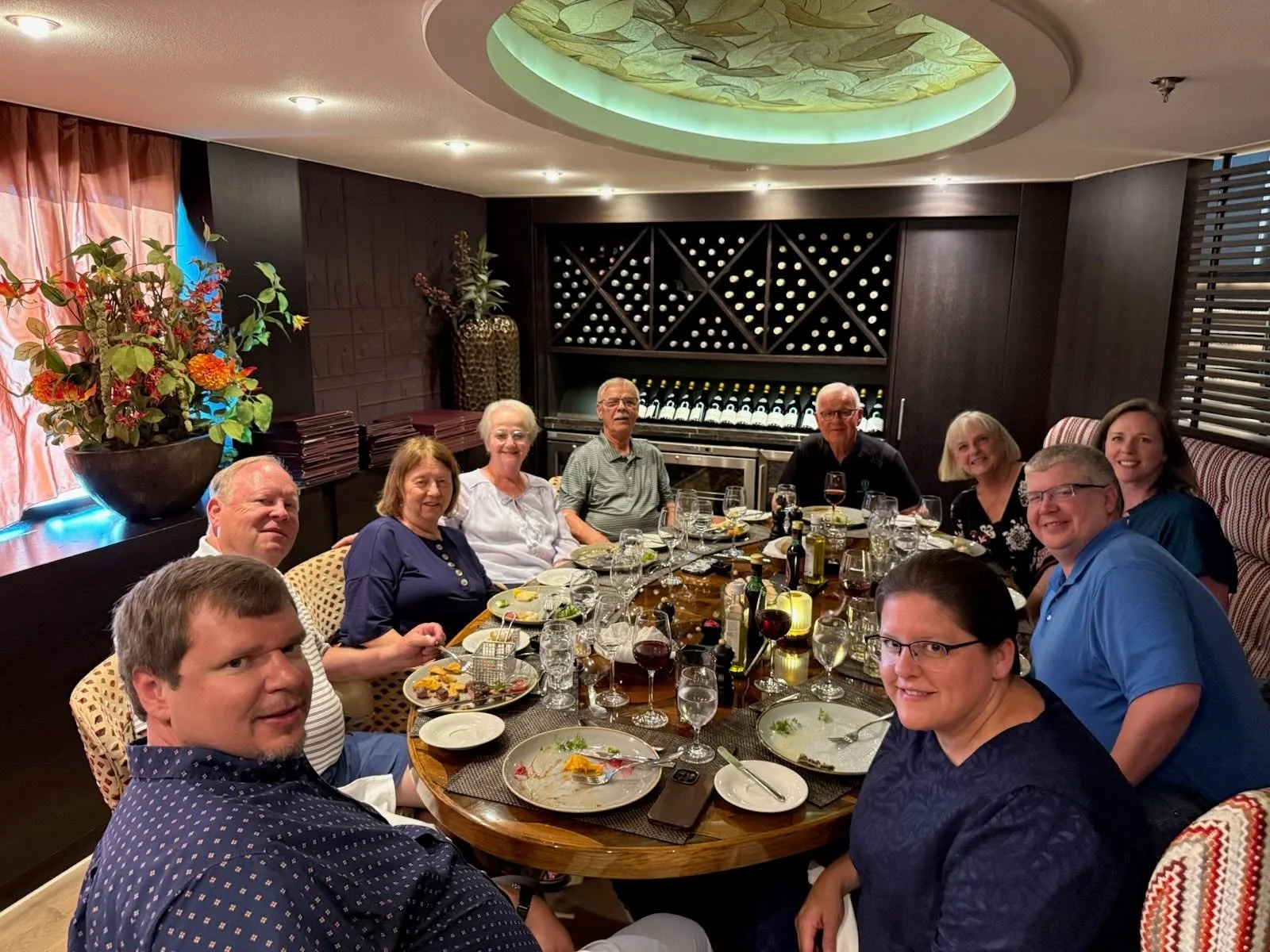 A group of ten people sitting around a dining table in a restaurant, smiling and enjoying a meal with wine glasses and plates of food, with dark wooden walls and a wine storage unit in the background.