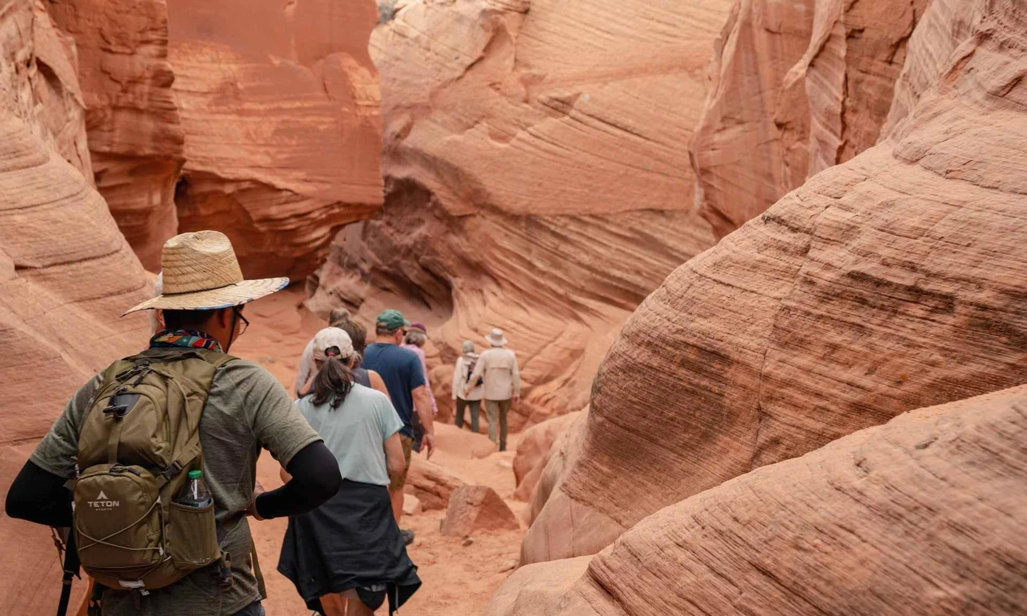 A group of hikers walking through a narrow canyon with red rock walls, some of the hikers wearing hats and backpacks.