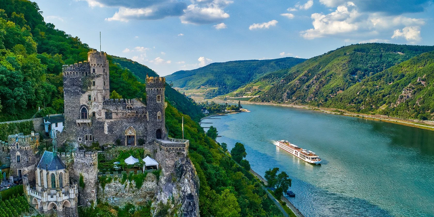 A castle situated on a hill overlooking a river with a boat cruising on it, surrounded by green mountains and a partly cloudy sky.