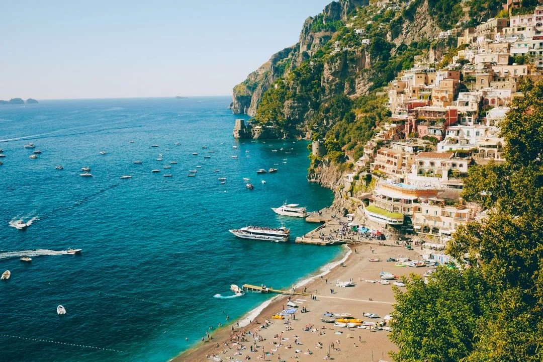 Coastal scene of Positano, Italy with colorful buildings on a hillside, a sandy beach, and boats in the water.