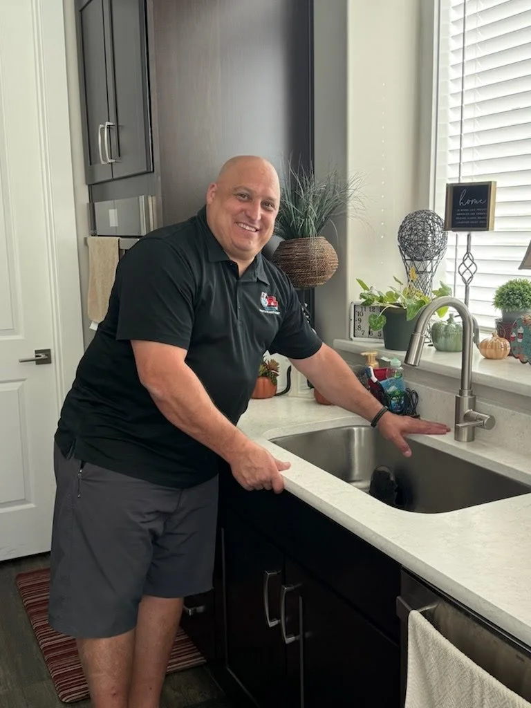 A man with a shaved head smiling and standing in a modern kitchen, leaning on a white countertop with a sink, surrounded by plants and decorative items.