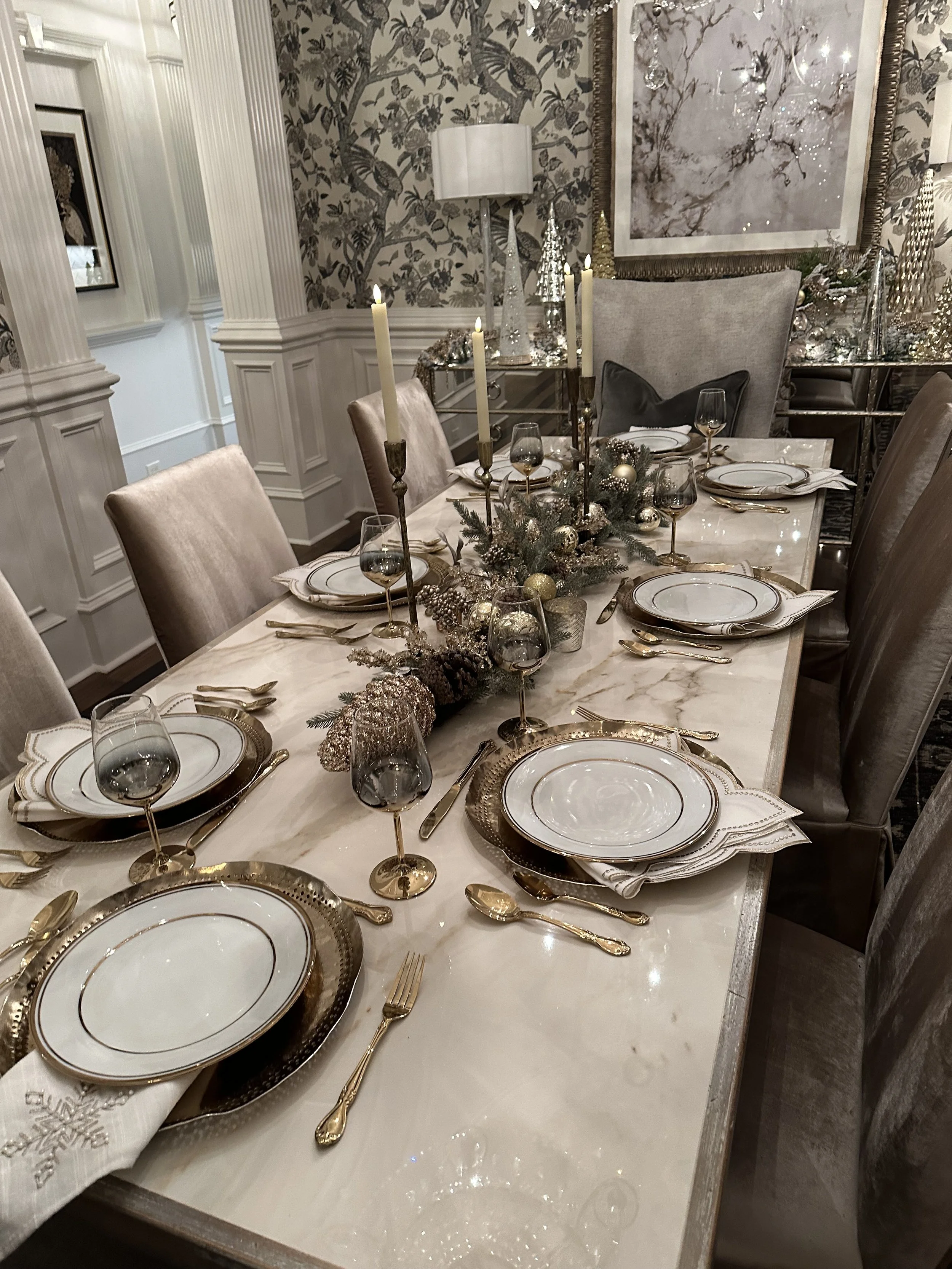 An elegant dining table set for a holiday dinner, decorated with gold and silver accents, candles, plates, glasses, and a centerpiece of pine branches and ornaments, with a modern textured wall and artwork in the background.