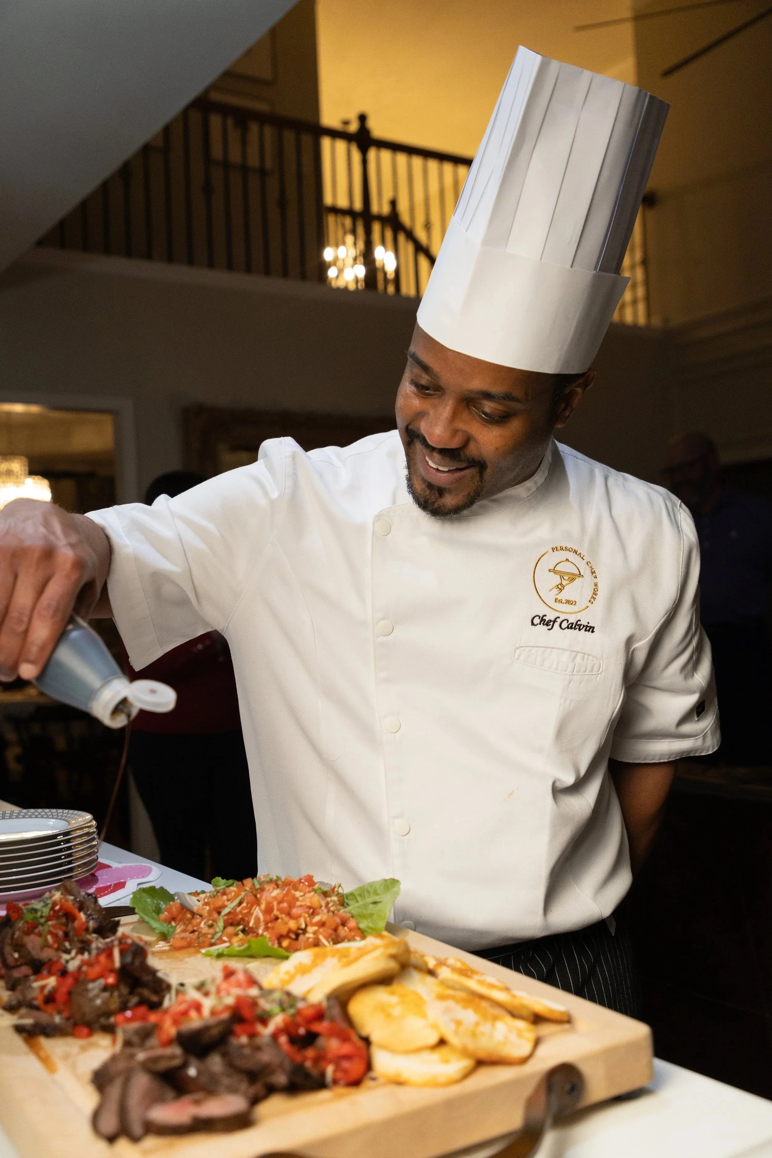 A chef in a white uniform and tall hat preparing food at a buffet. There are various dishes, including sliced fruit and salads, on a wooden serving board in the foreground.