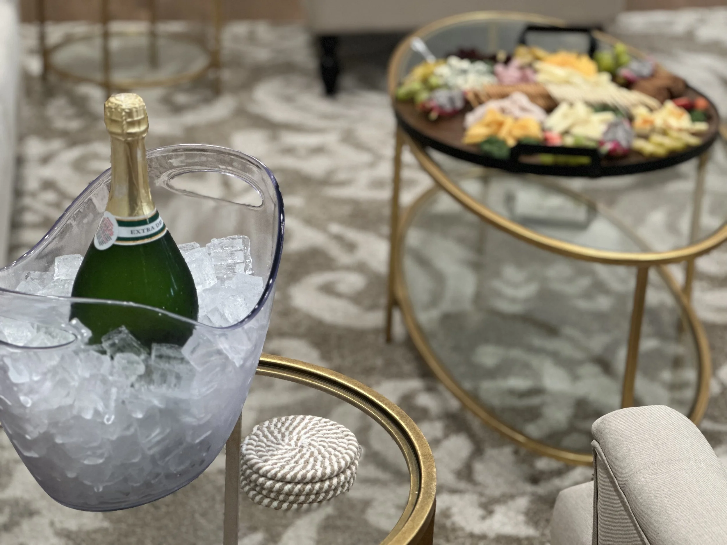 A bottle of sparkling wine in an ice bucket in the foreground. In the background, a round coffee table with a large snack platter containing assorted cheeses, meats, and vegetables.