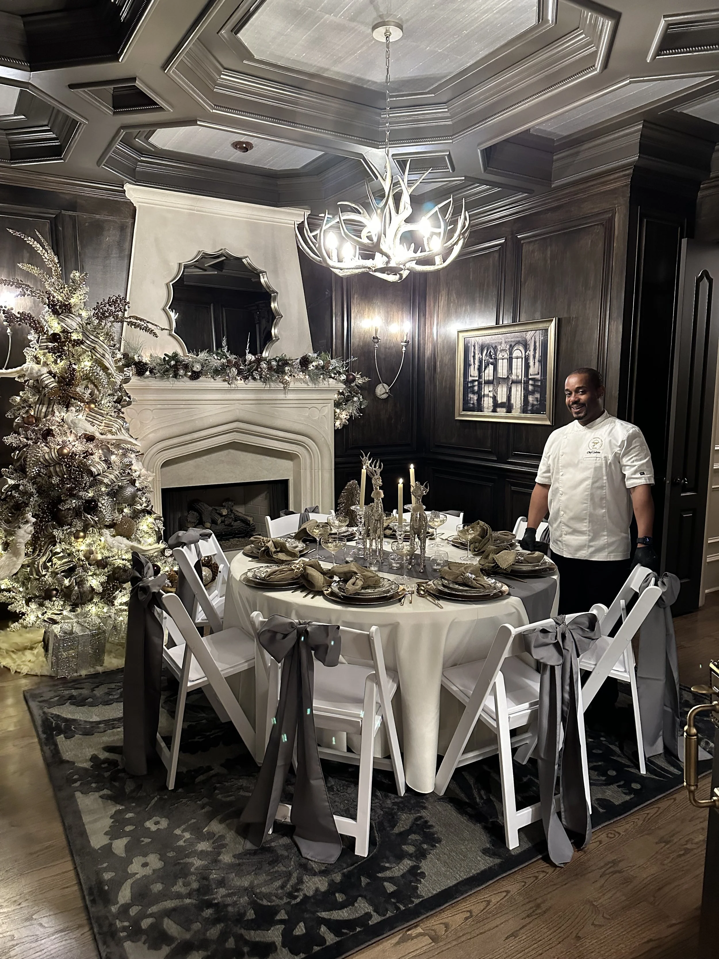 A luxurious dining room decorated with a Christmas theme, featuring a round table set with gold and beige napkins, a Christmas tree with white and gold ornaments, a white fireplace with a festive garland, dark wood-paneled walls, a chandelier, and a man in a white chef's coat standing near the table.