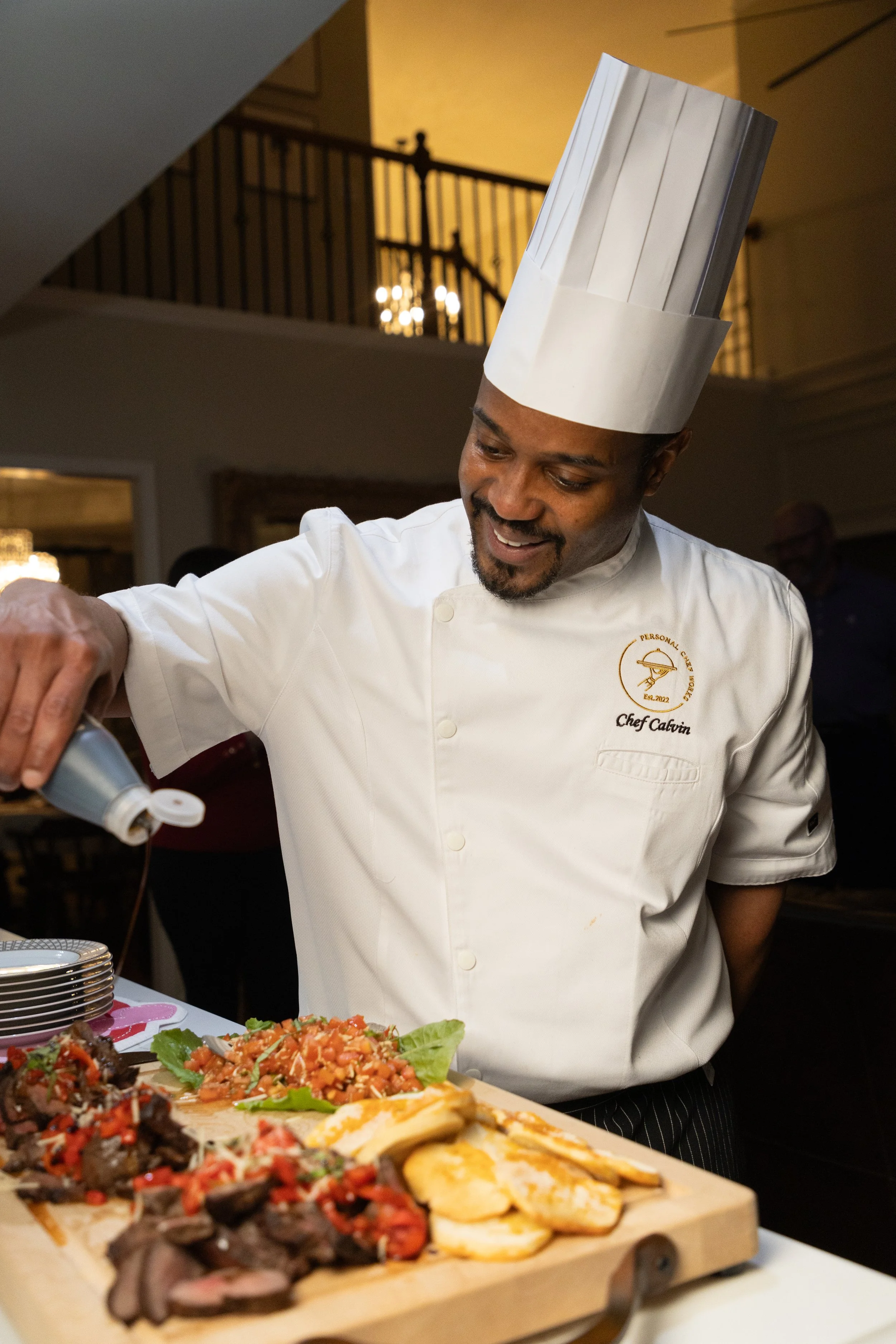 A chef in a white uniform and tall hat is smiling and serving food at a buffet. The buffet includes sliced meats, grilled vegetables, and salad. There are plates stacked on the side. The background shows a staircase and other people in the restaurant.