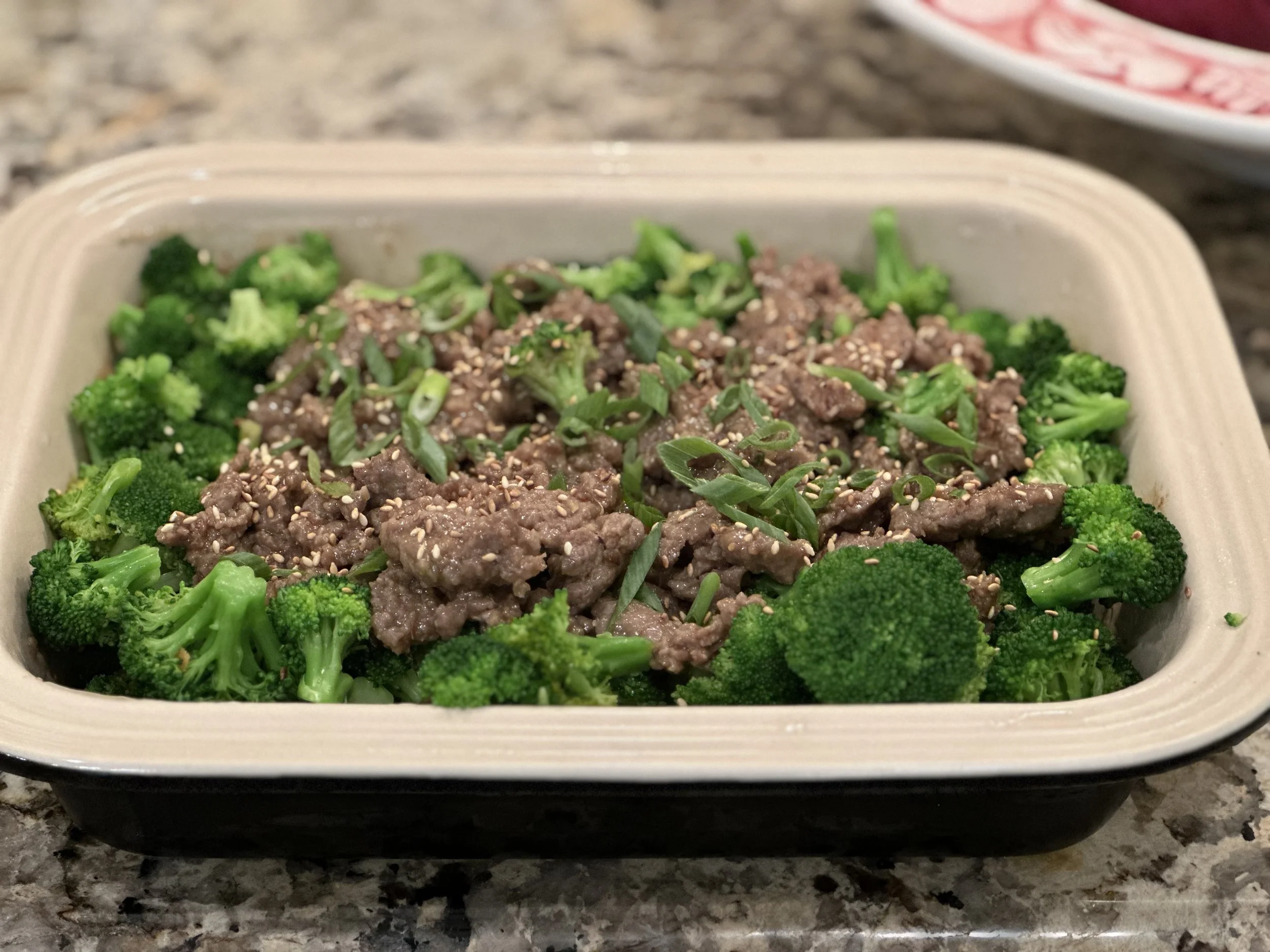 A meal of cooked broccoli florets topped with ground beef, chopped green onions, and sesame seeds in a beige container on a kitchen countertop.