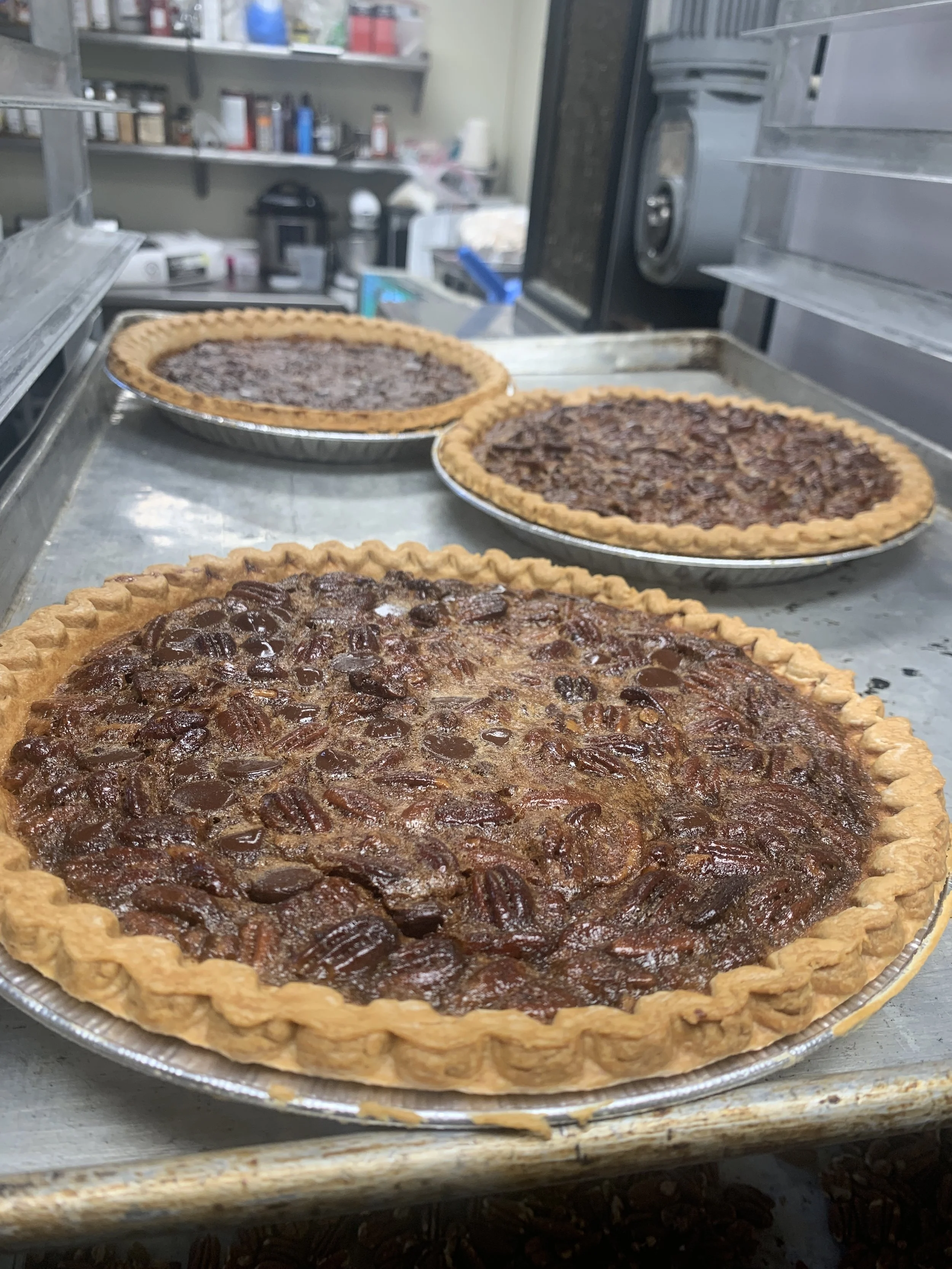 Close-up of three pecan pies with golden crusts on a metal tray, in a bakery or kitchen setting.