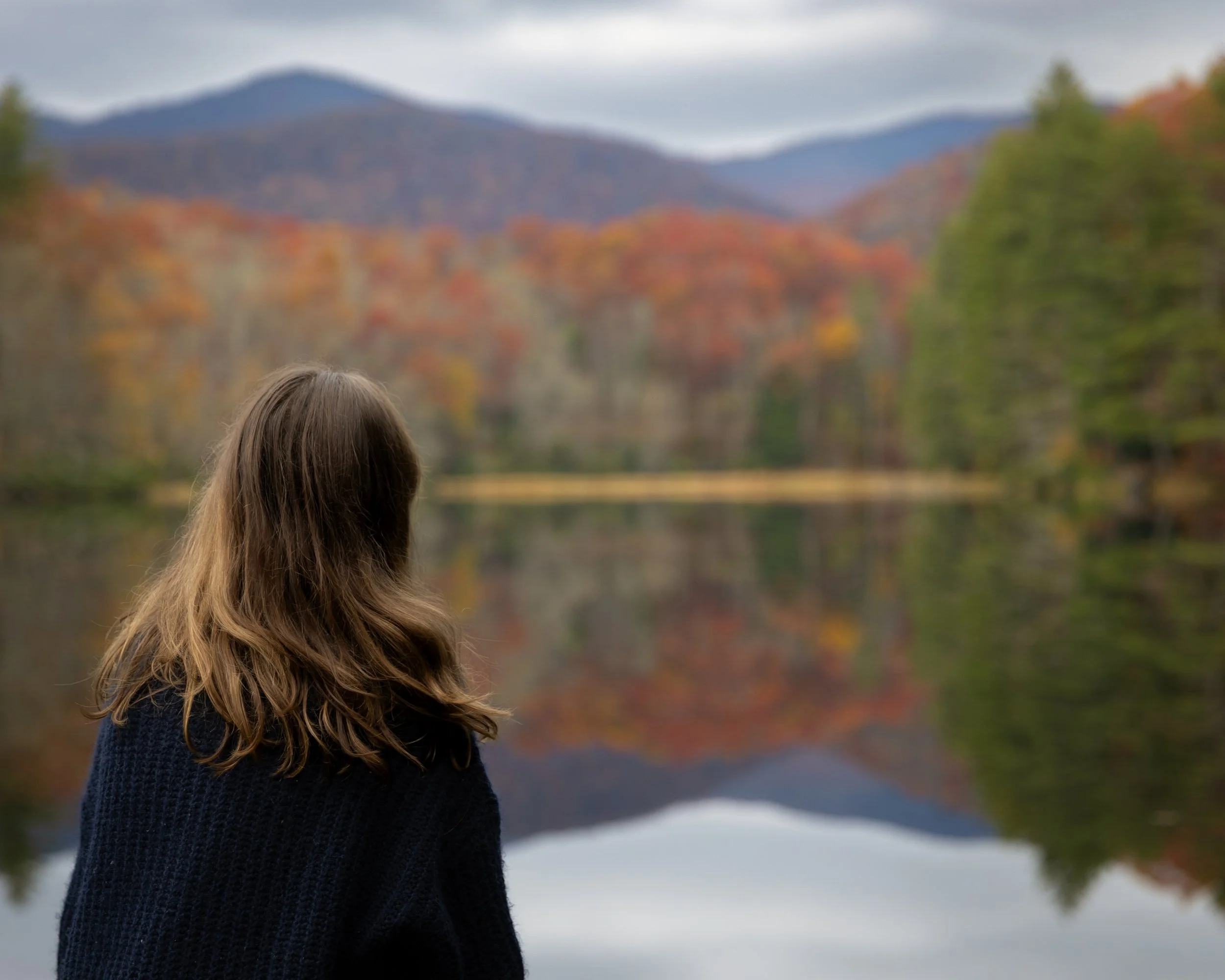 A woman with long brown hair, wearing a dark blue sweater, stands outdoors near a lake or river with a mountain and colorful trees in the background during fall.