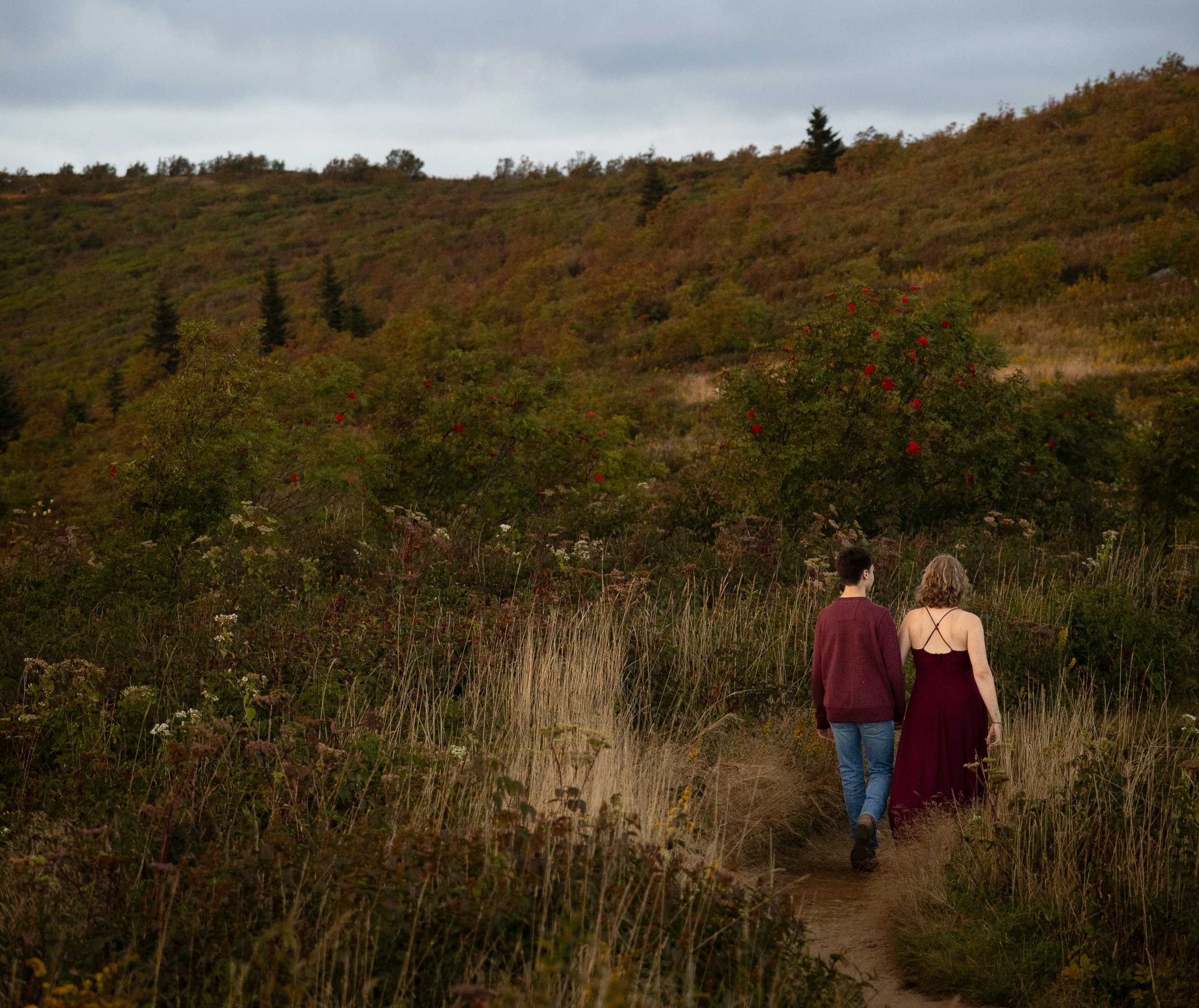 A man and woman walking hand-in-hand along a dirt trail in a hilly, enclosed landscape with greenery, shrubs, small flowers, and scattered trees, under a cloudy sky.
